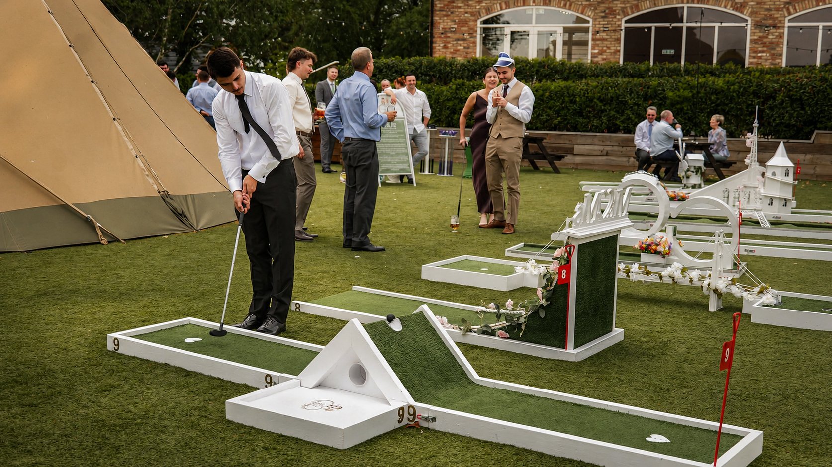 People playing mini golf outdoors at a social event, with a tent on the left, greenery, and a brick building in the background.