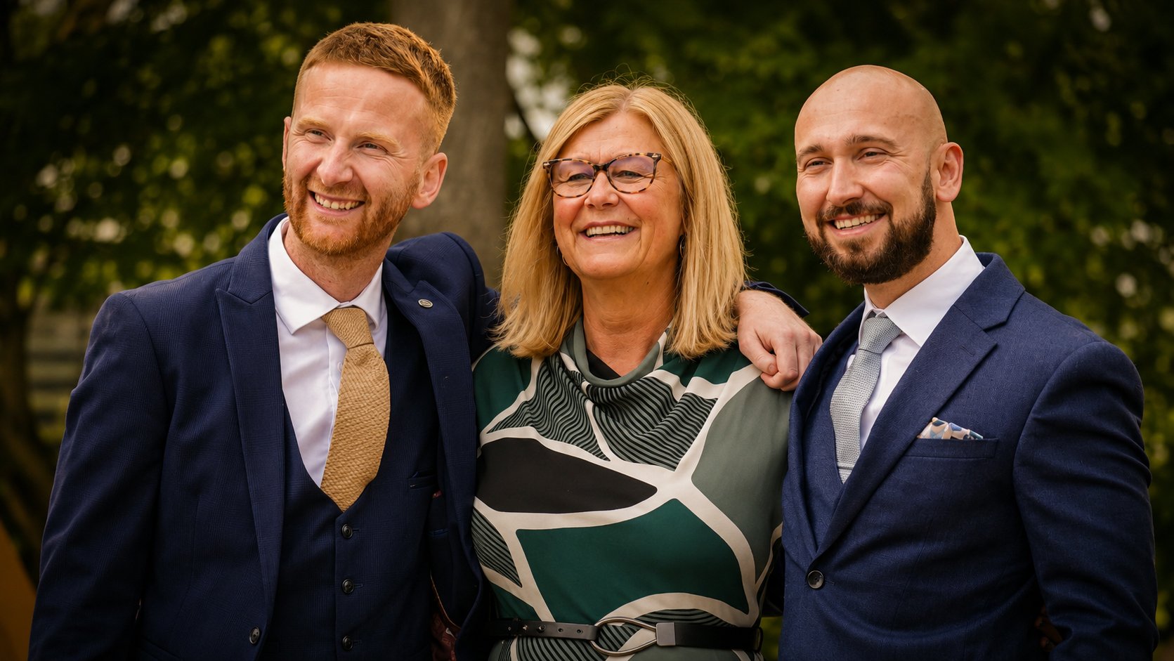Three adults, two men and one woman, smiling and posing outdoors in formal attire, with greenery in the background.