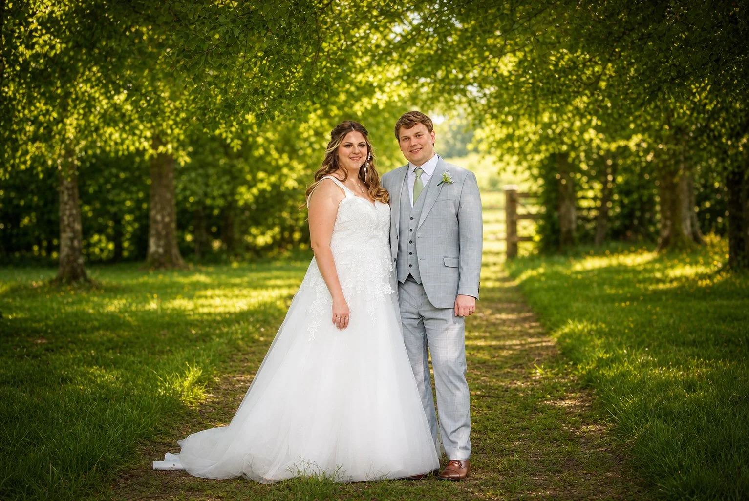 A bride and groom standing together outdoors in a grassy area with trees in the background, on their wedding day.