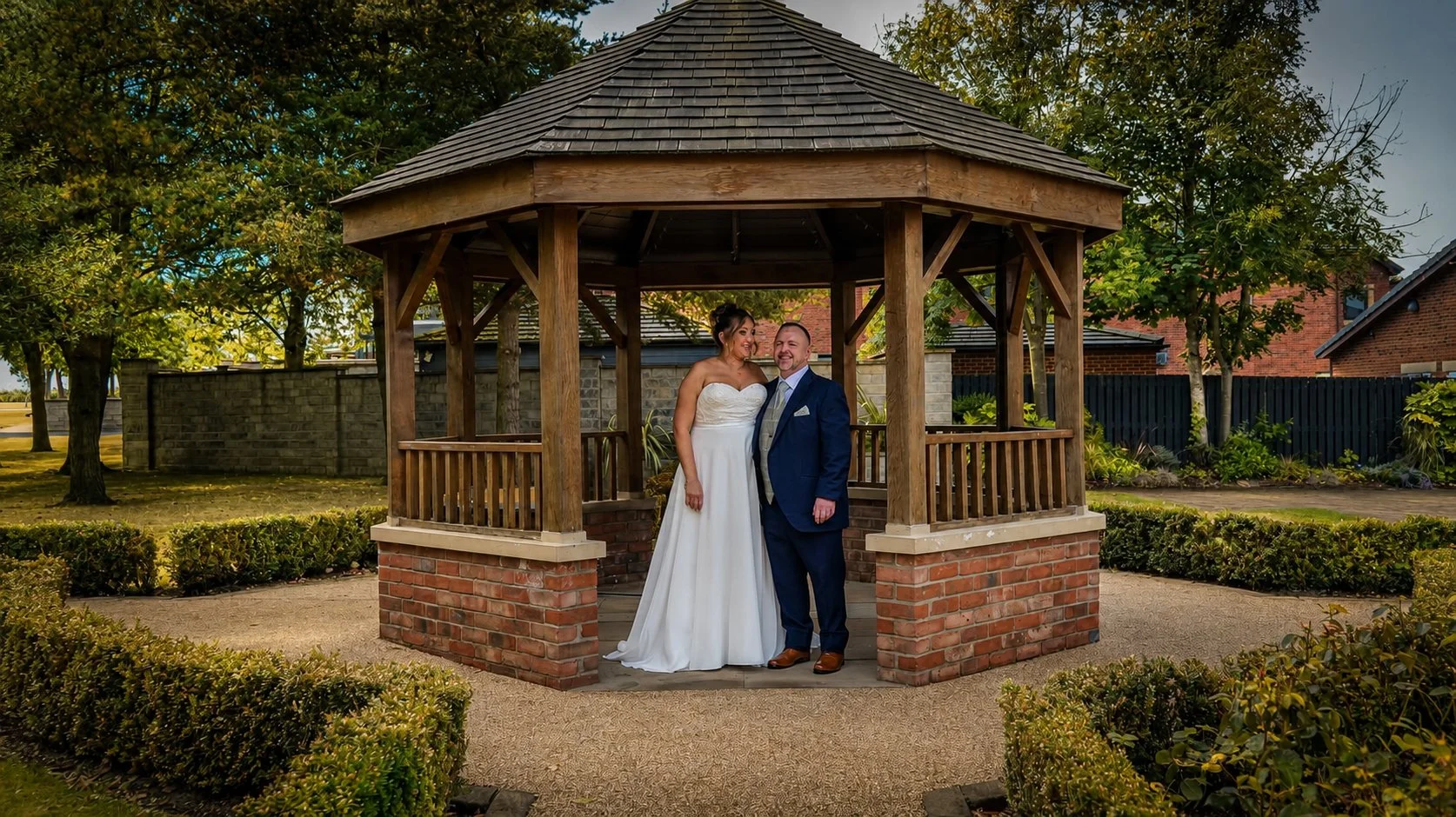 A bride and groom standing together in a wooden gazebo with a brick base, celebrating a wedding outdoors during daytime. The bride is wearing a white strapless wedding dress, and the groom is dressed in a navy suit with a grey tie. They are smiling a
