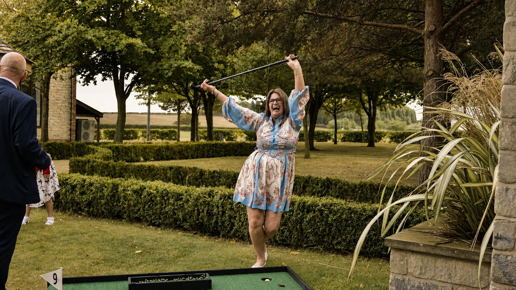 Woman celebrating joyfully outdoors, holding a golf club in the air, with other people in the background on a golf course or garden setting with trees and bushes.