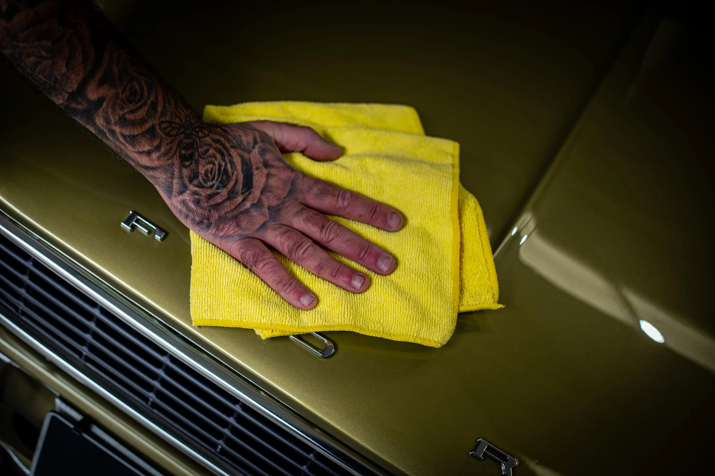 A person's hand with tattoos is holding a yellow microfiber cloth and cleaning the hood of a metallic gold car.
