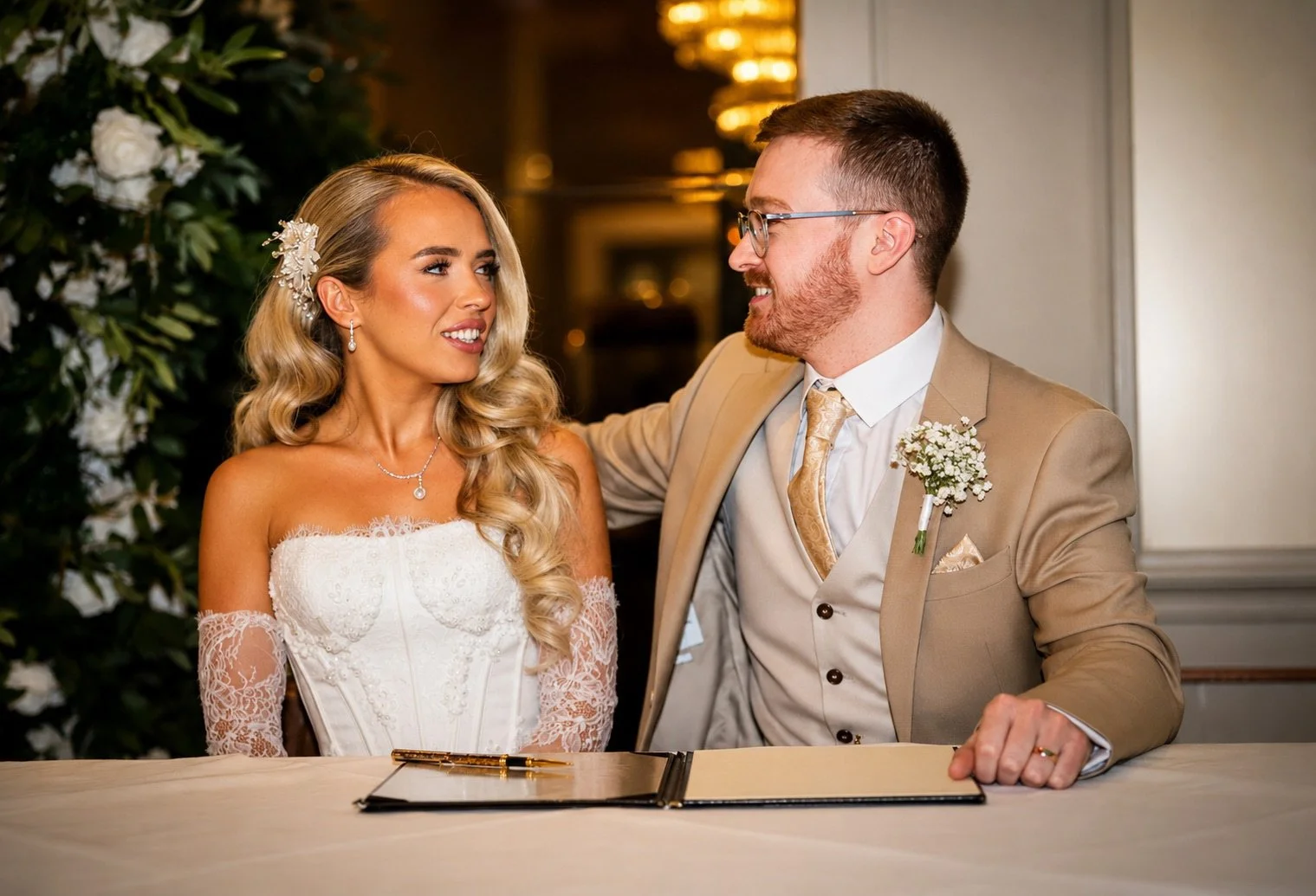 A bride and groom sitting at a table, smiling at each other, during their wedding ceremony. The bride has long blonde hair, wearing a white lace dress and jewelry. The groom has short hair, glasses, a beige suit with a boutonniere, and a gold tie. Th