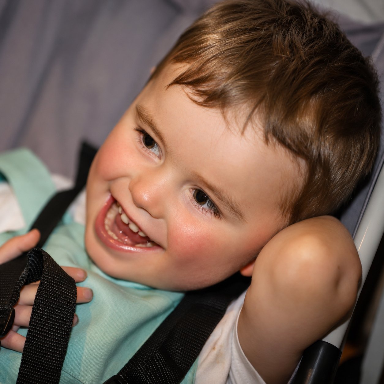 A young boy with brown hair smiling and lying in a stroller with his hand behind his head.