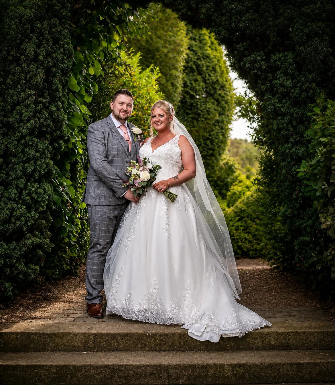 A bride and groom standing together outdoors in a lush green garden, smiling, with the bride holding a bouquet of flowers.