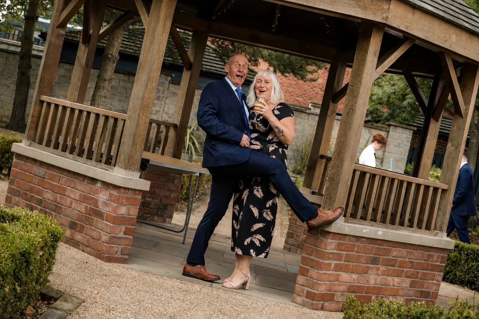 A man in a dark suit and a woman in a floral dress laughing and posing together under a wooden gazebo in a garden.
