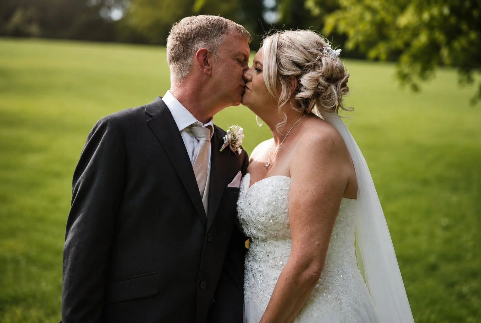 A bride and groom share a kiss outdoors on a grassy field with trees in the background, during a wedding celebration.