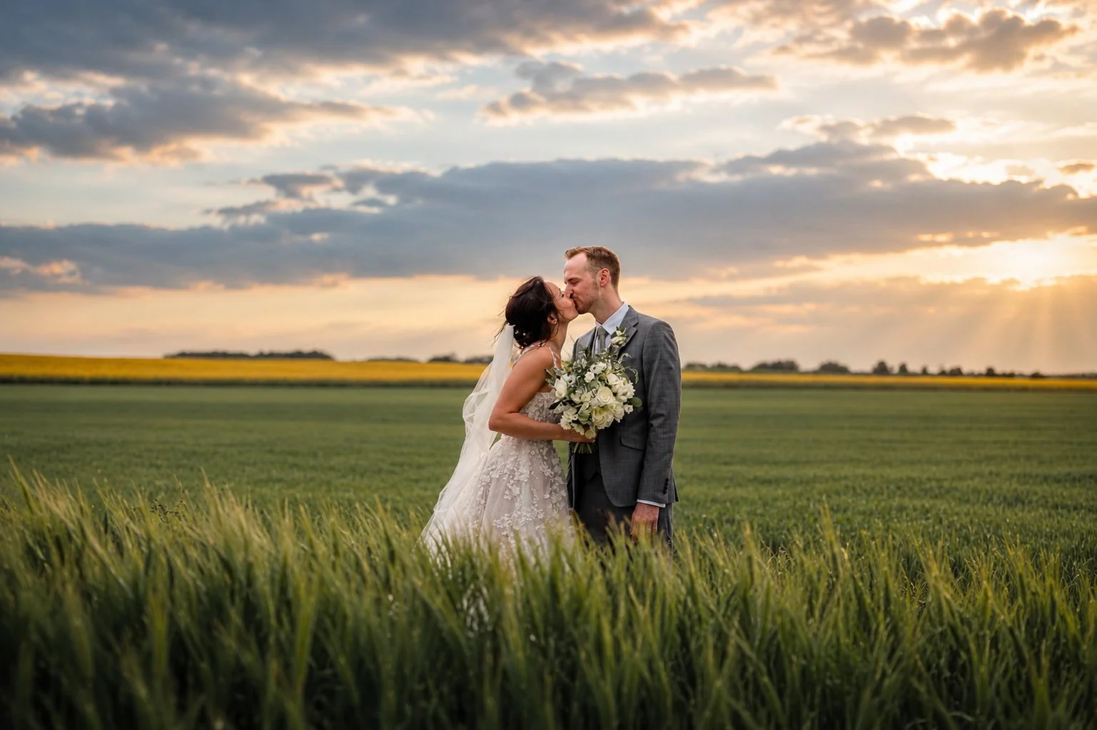 A bride and groom kiss in a grassy field at sunset, with the bride holding a bouquet of white flowers, the sky filled with clouds and the sun rays shining through.