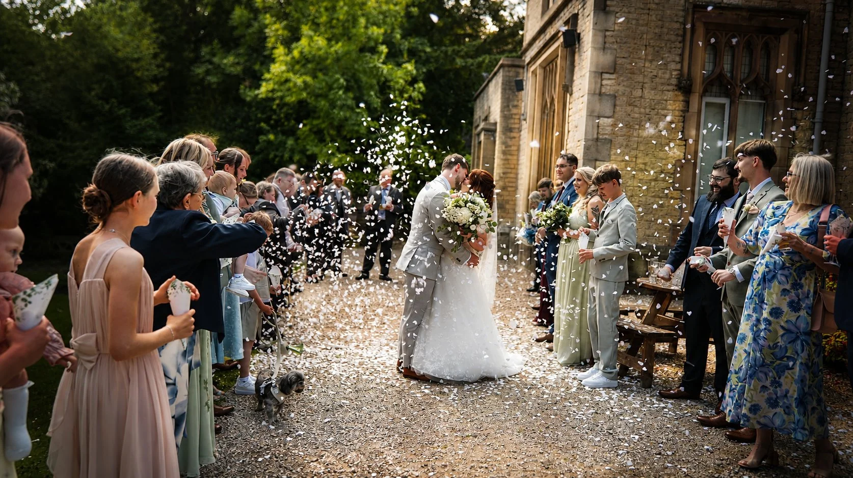 Wedding ceremony outdoors with bride and groom kissing amidst guests throwing confetti.