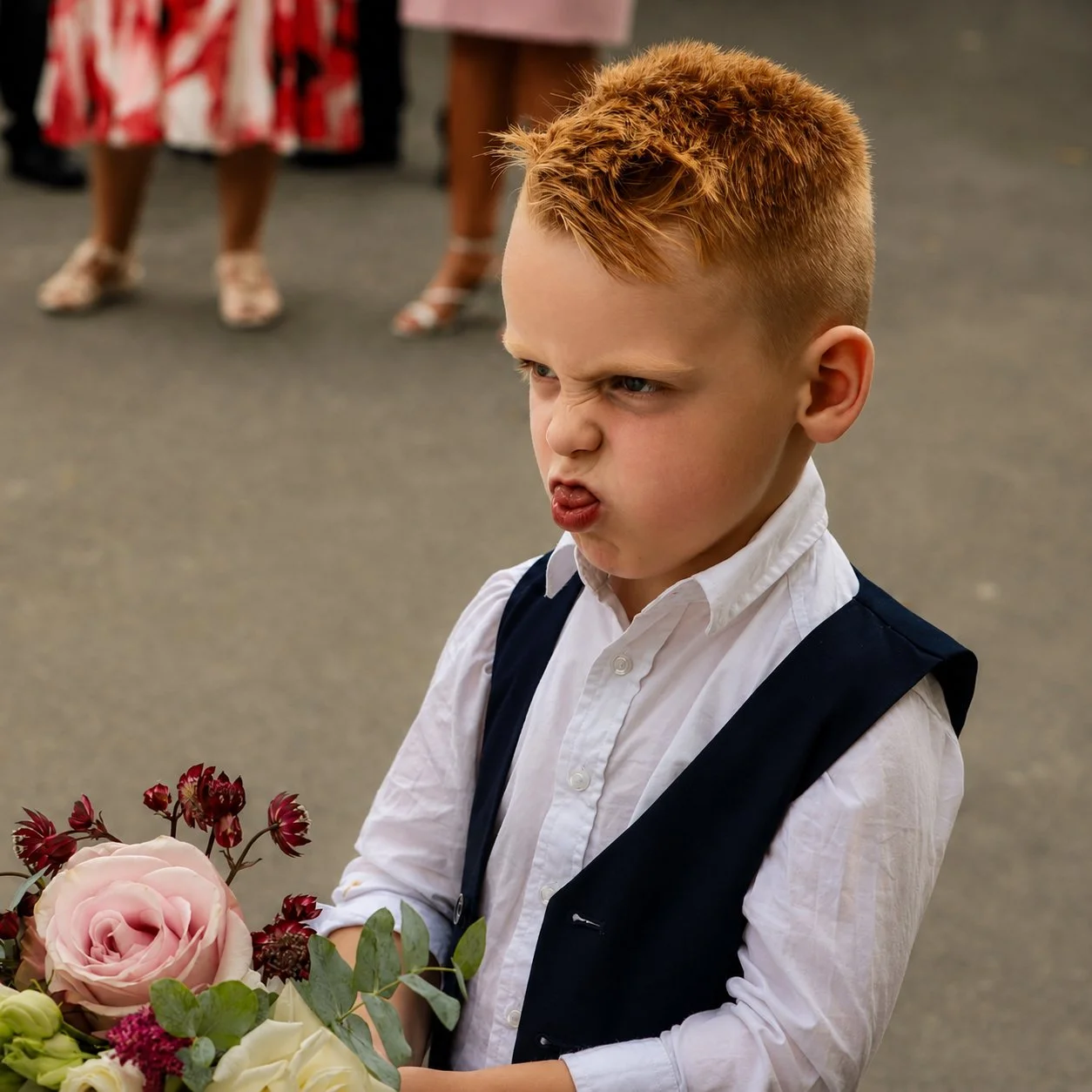 A young boy with reddish hair making a funny face, holding a bouquet of flowers, at what appears to be a formal or special event.
