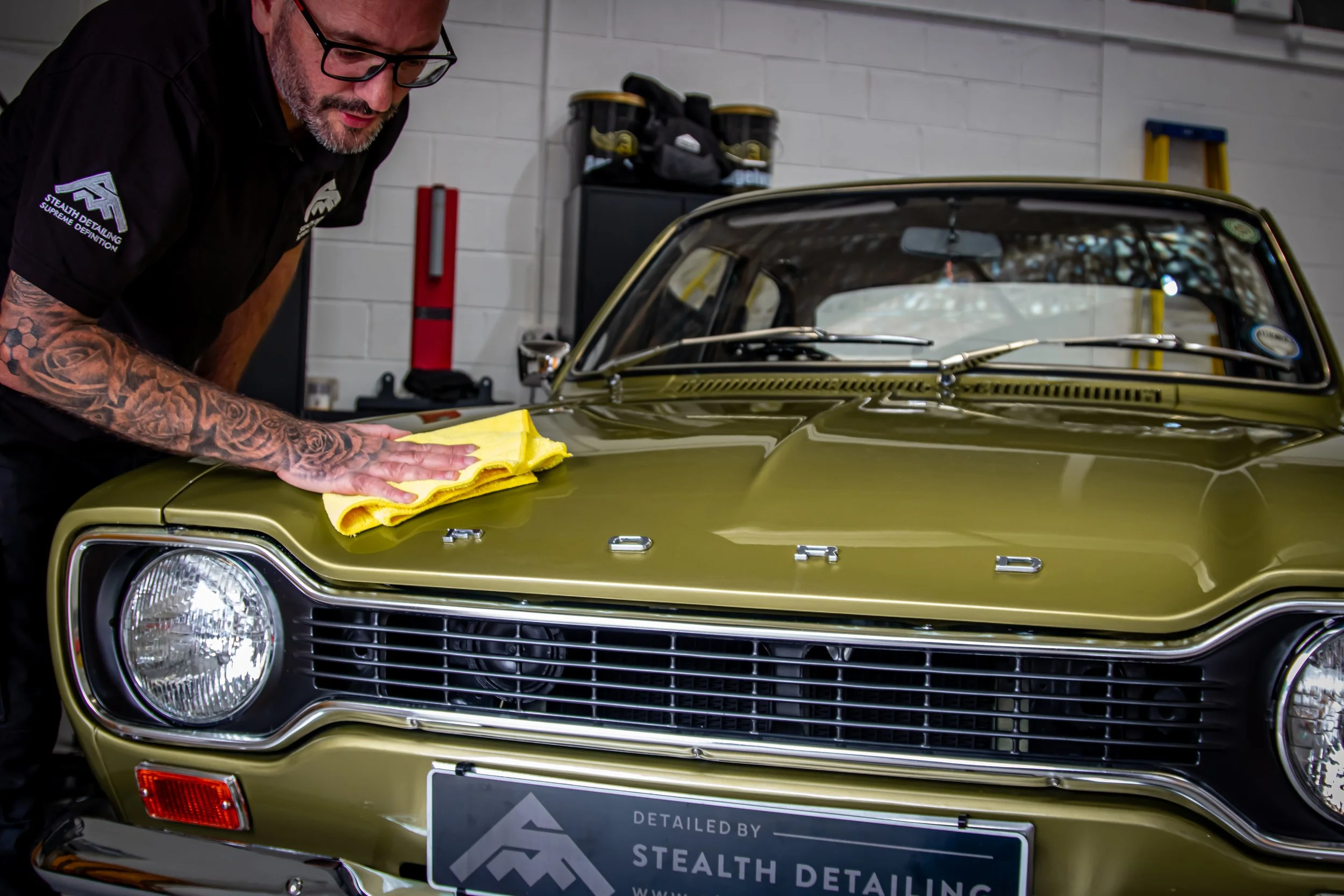 A man with tattoos on his right arm is polishing the front of a vintage gold-colored car inside a garage. He is wearing glasses and a black shirt with a logo on the sleeve. The garage has a white brick wall background with tools and equipment. The ca