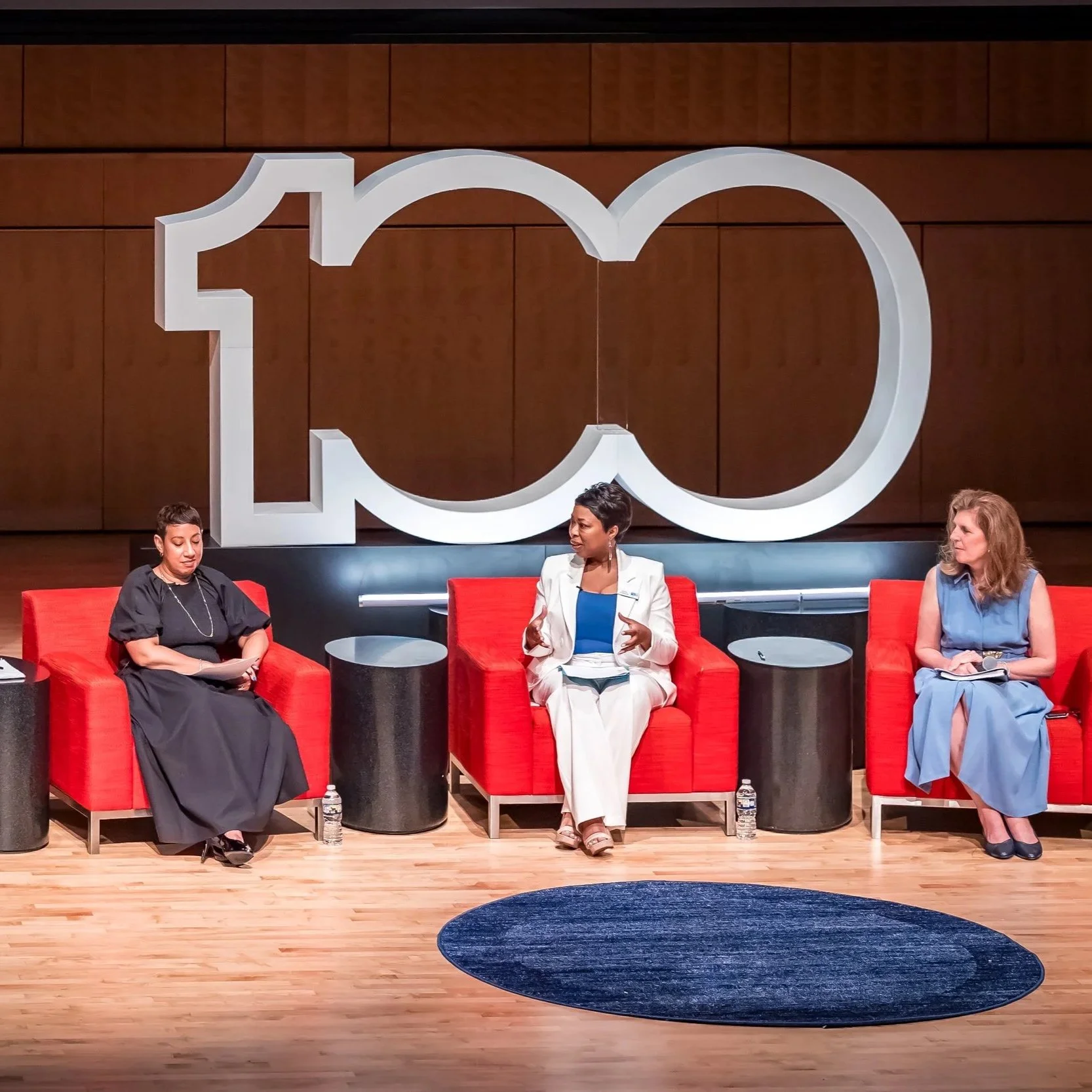 Three women sitting on stage during a conference panel discussion with a large white dimensional logo backdrop.