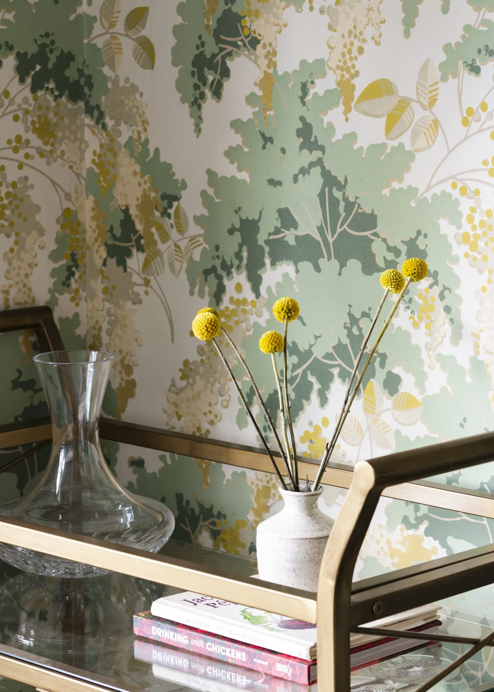 Decorative glass table with a white vase holding yellow spherical flowers, a clear glass decanter, and a stack of books against a wall with botanical patterned wallpaper.