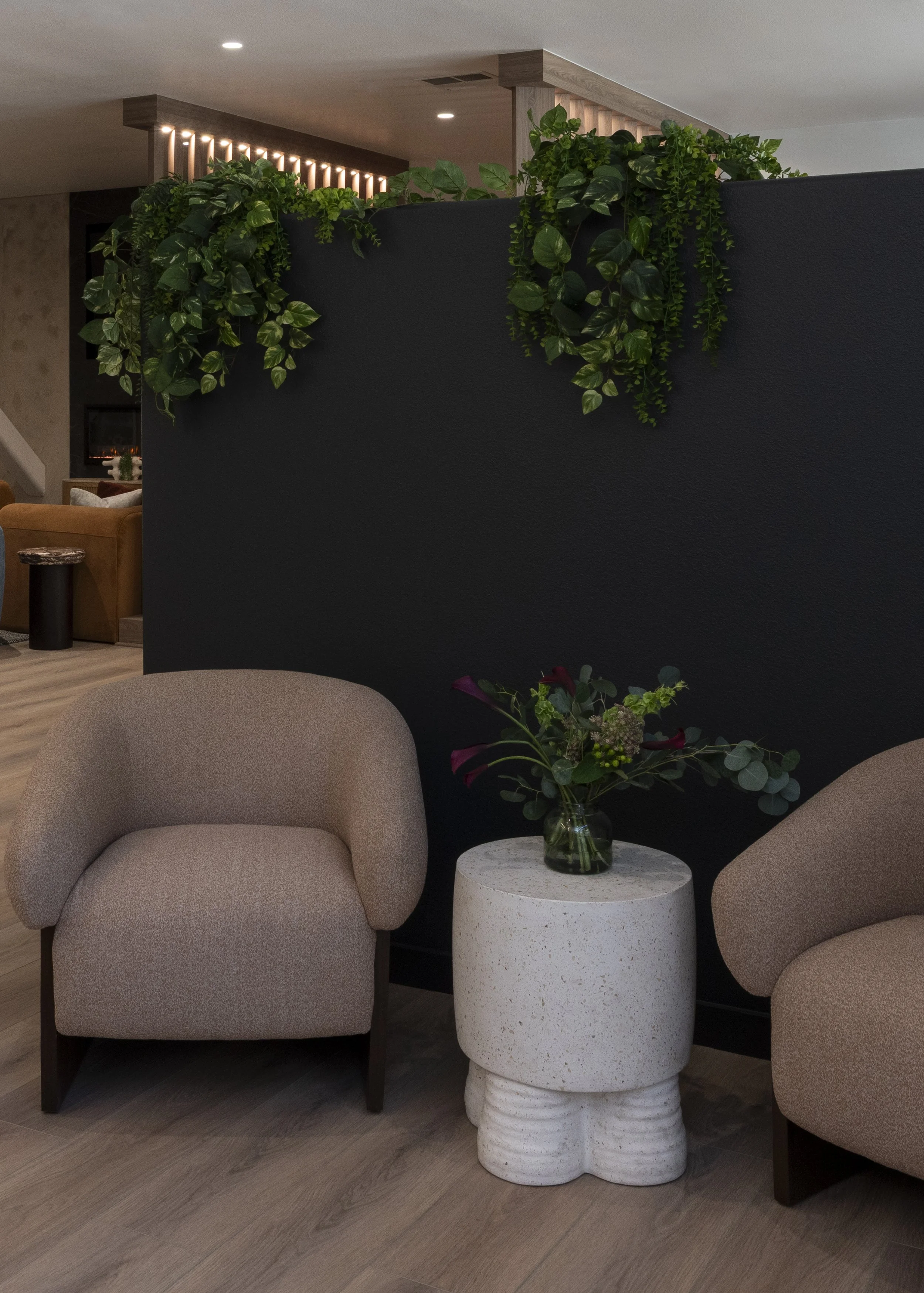 A cozy indoor seating area with two beige armchairs, a white textured side table with a flower vase, and green plants on a black partition wall.