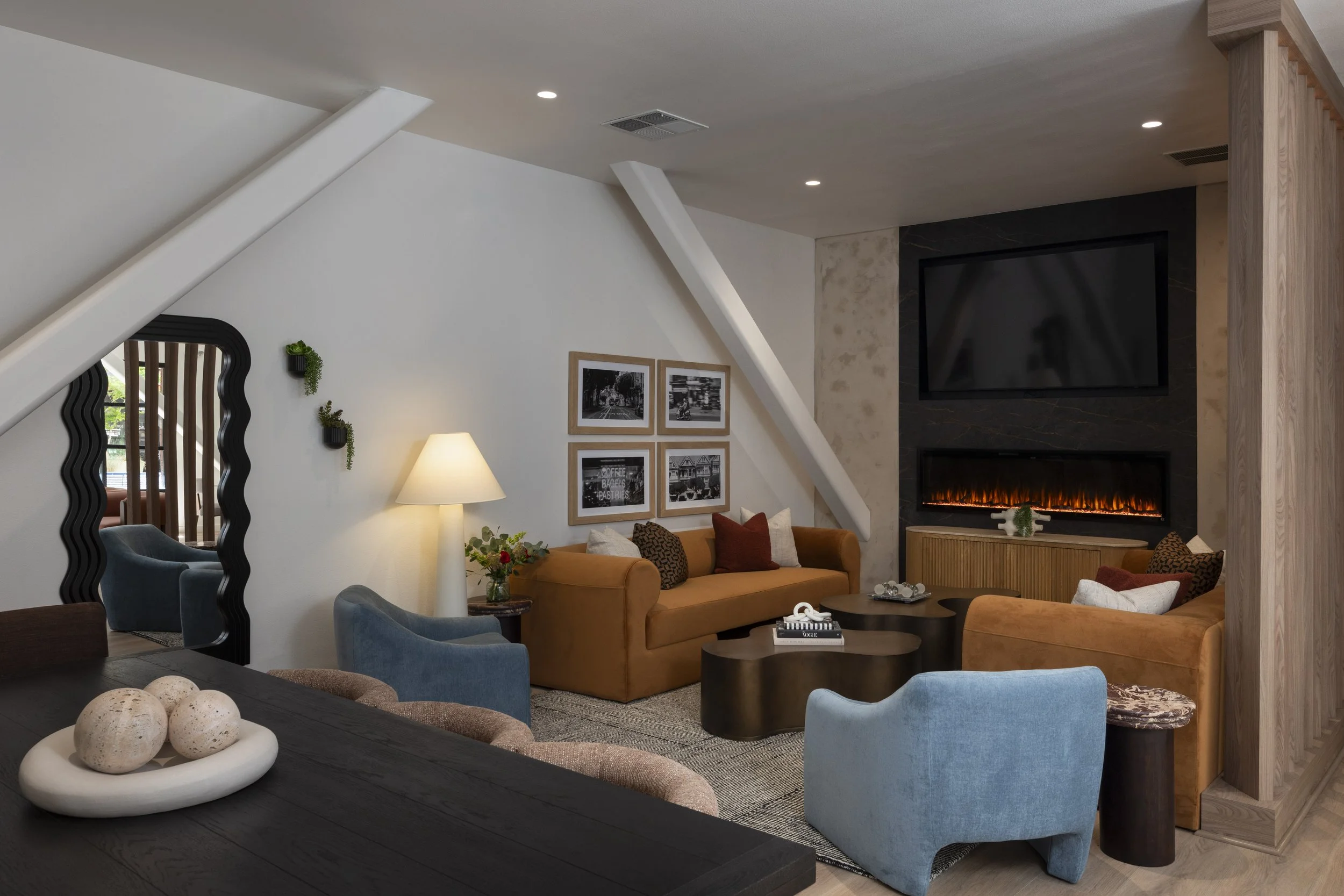 Living room with modern decor, featuring a mustard yellow sofa, light blue and brown chairs, a black coffee table, wall art, a wall-mounted TV, and a fireplace with flames.
