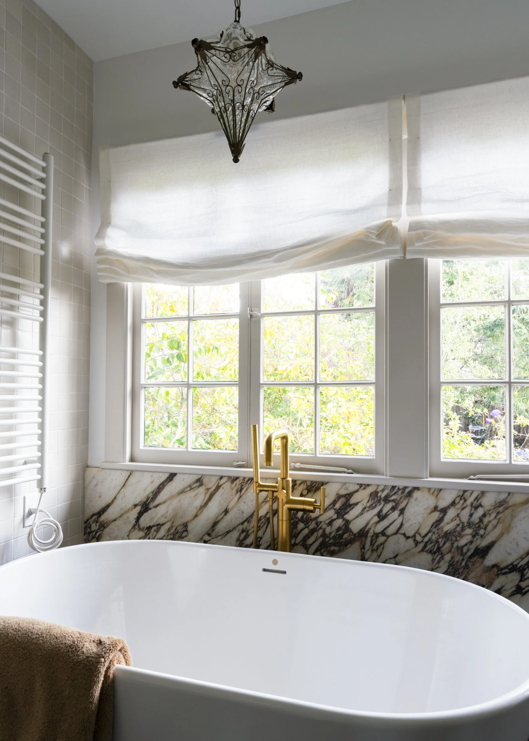 A bathroom with a white freestanding bathtub, a large window with white windowpanes, and a marble backsplash. There is a gold faucet on the bathtub, a brown towel hanging on the edge, and a decorative chandelier hanging from the ceiling.