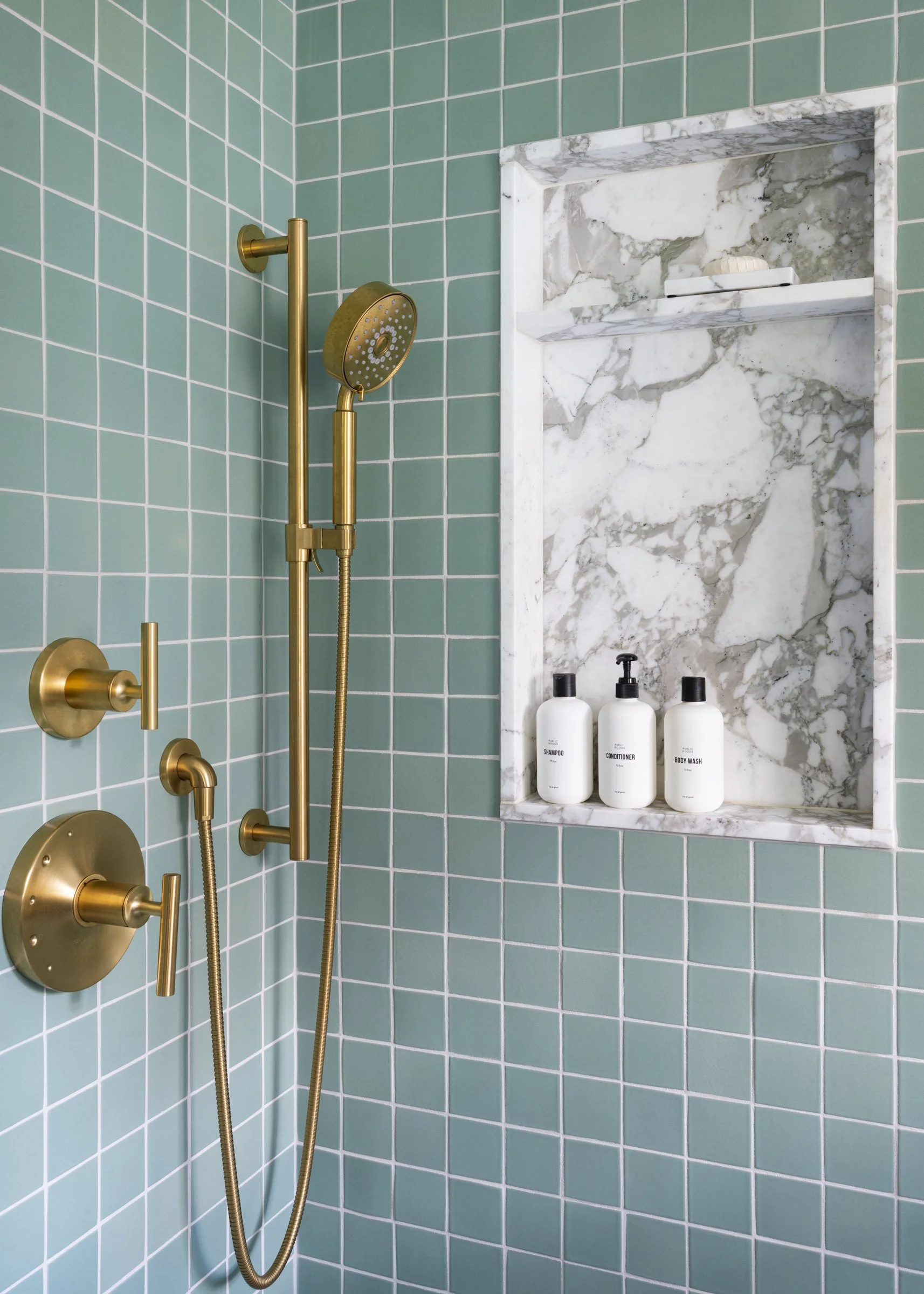 A shower with teal square tiles on the walls, gold fixtures including a showerhead and control handles, and a marble recessed shelf holding three white bottles labeled shampoo, conditioner, and body wash.