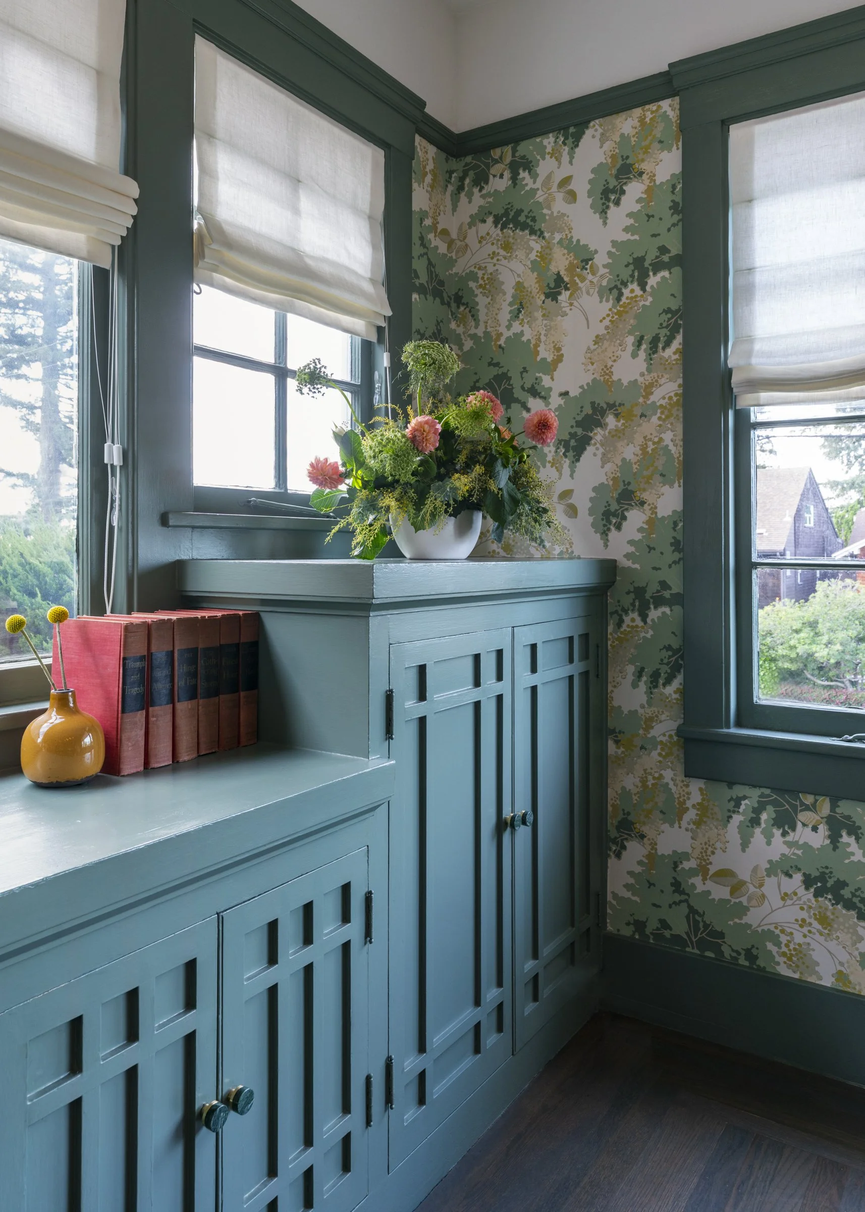 Corner of a room with teal built-in cabinets, floral wallpaper, two windows with white blinds, a plant with pink flowers on top, red books, and a small yellow vase.