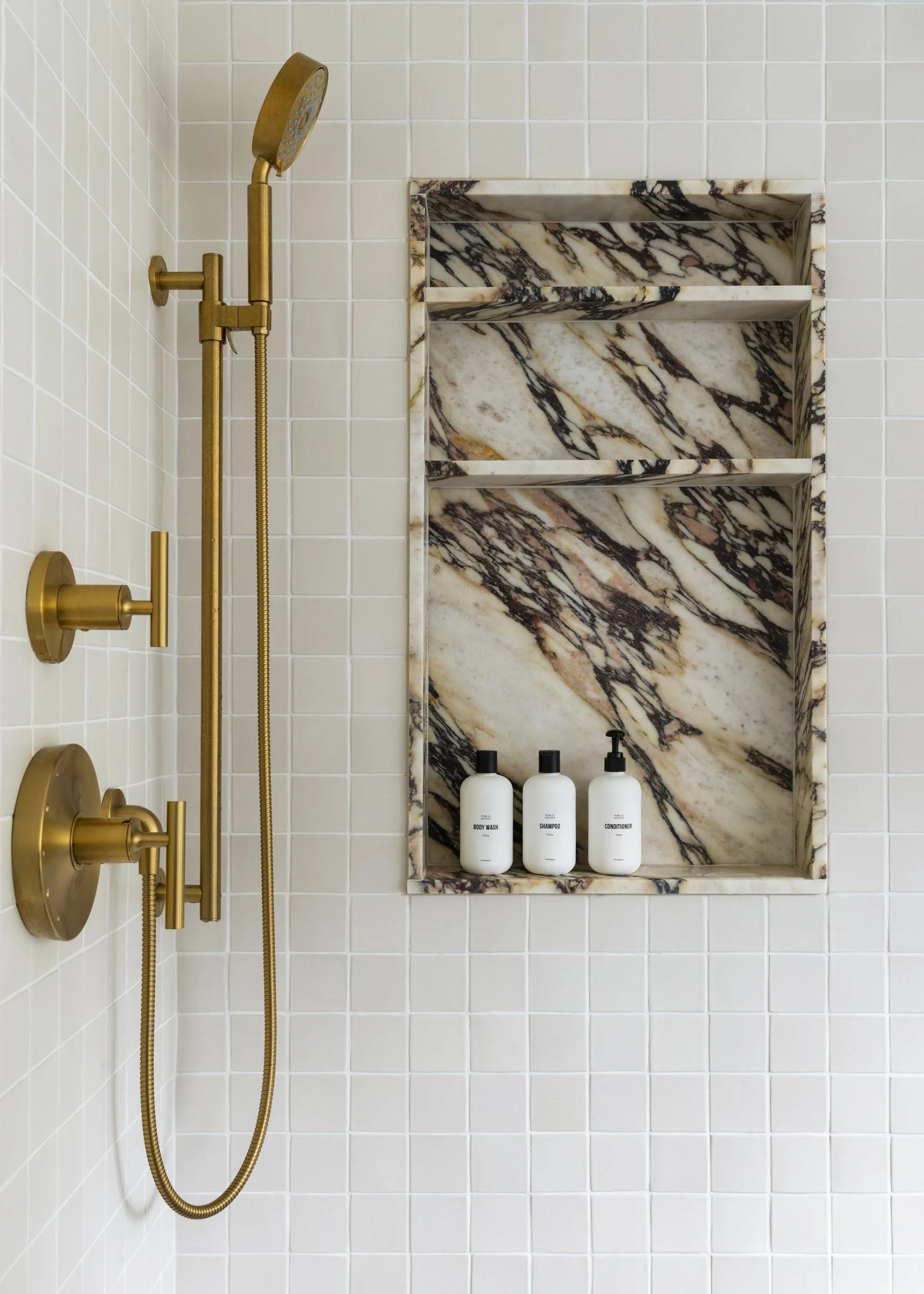 A shower with white tiled walls, a built-in marble niche with black and brown veining, and three white bottles labeled body wash, shampoo, and conditioner.
