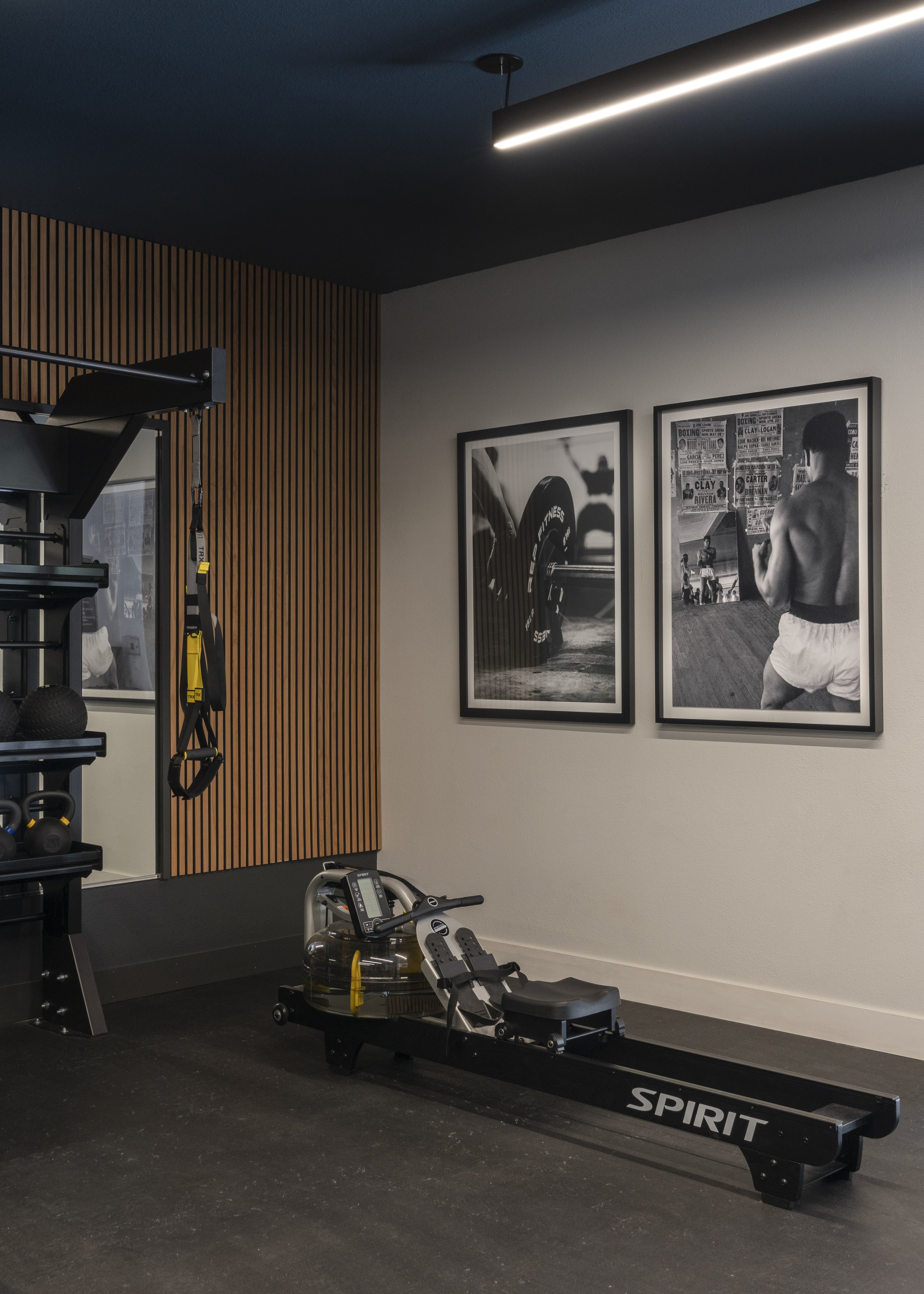A modern gym corner with a rowing machine, black and white fitness photographs on the wall, and workout equipment including kettlebells and resistance bands. Wood paneling decorates the wall.