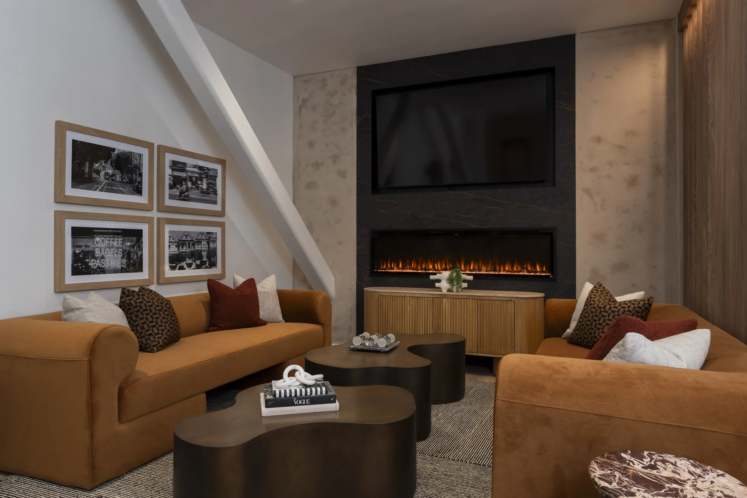 Modern living room with two tan sofas, black and white framed photos on the wall, a fireplace with a television above, next to a wooden cabinet, and a coffee table with decor items.