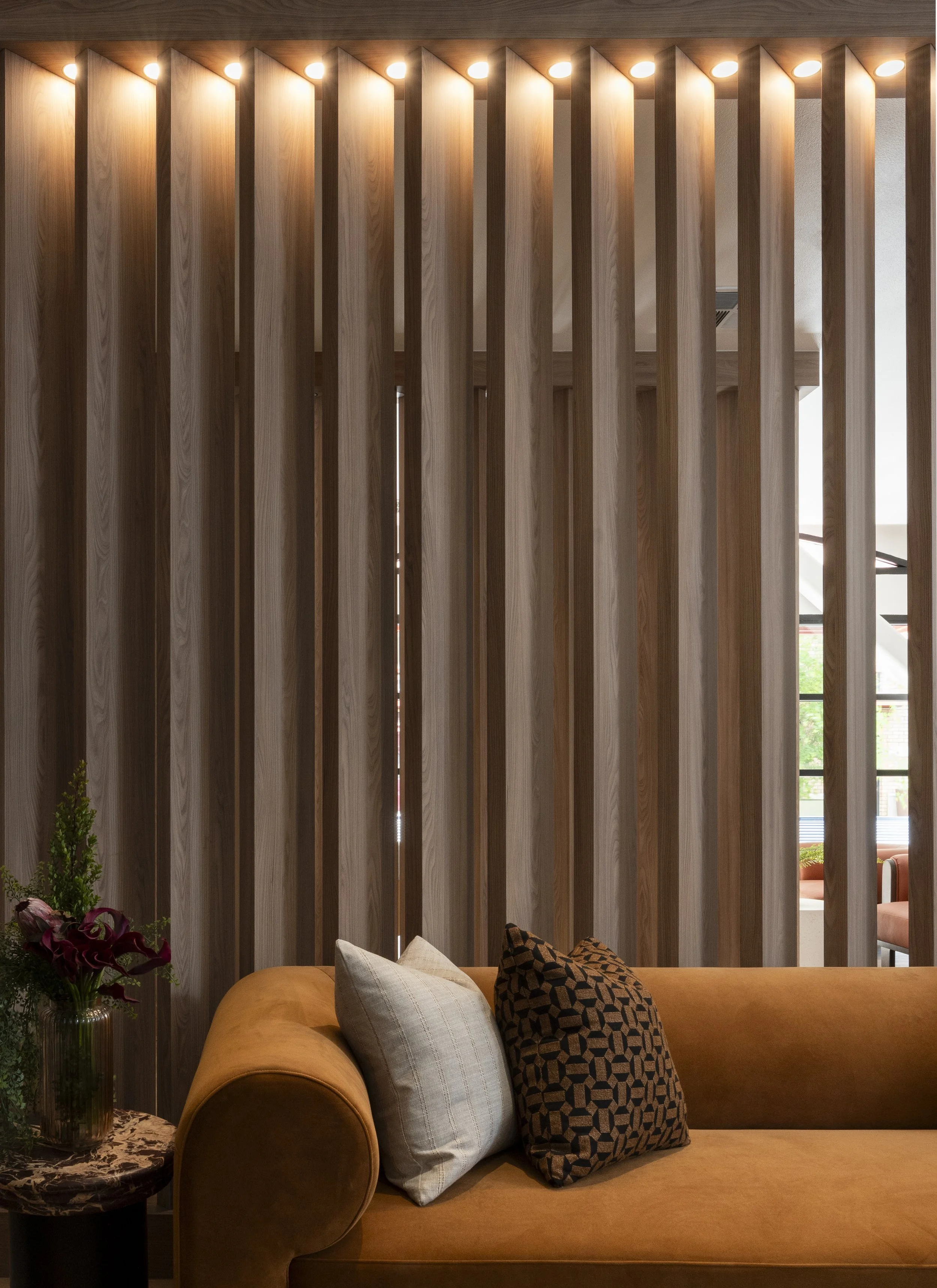 Interior of a modern room with a mustard-colored sofa, throw pillows, a side table with flowers, and wooden vertical slats with lights on top.