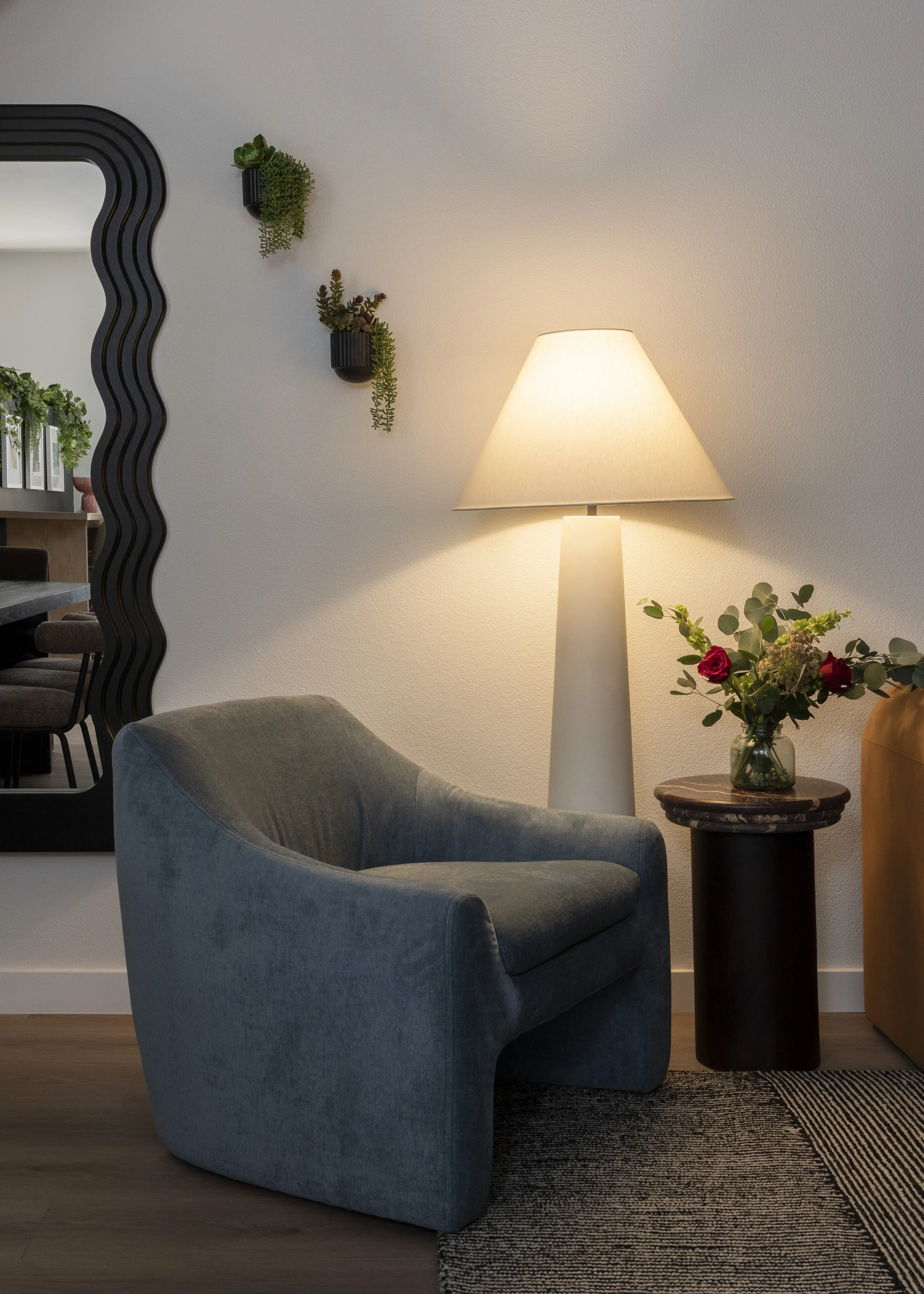 Living room corner with gray armchair, white floor lamp, round side table with a vase of flowers, and a wall mirror.