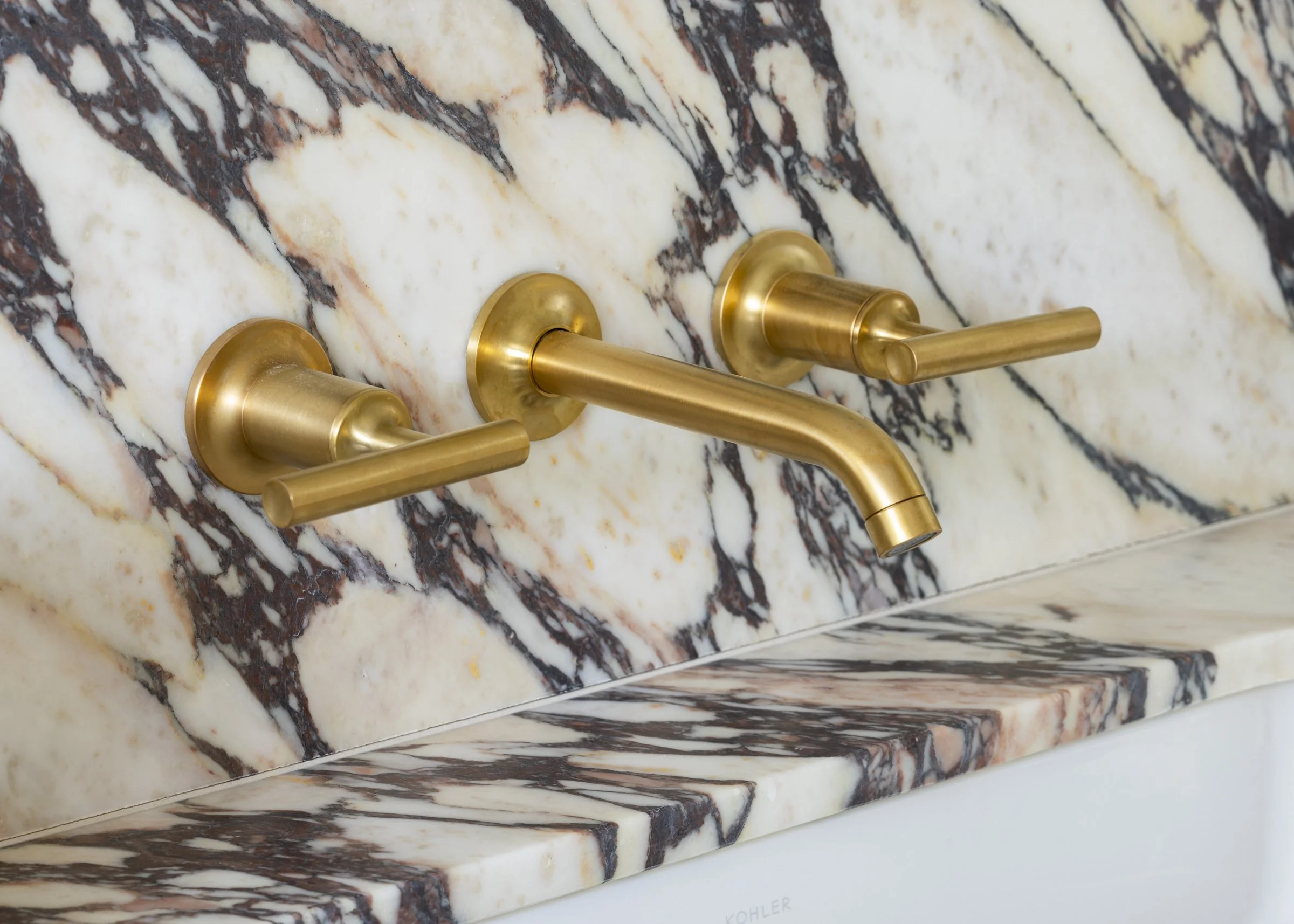 A wall-mounted gold faucet with two handles on a marble backsplash with dark and light veins.