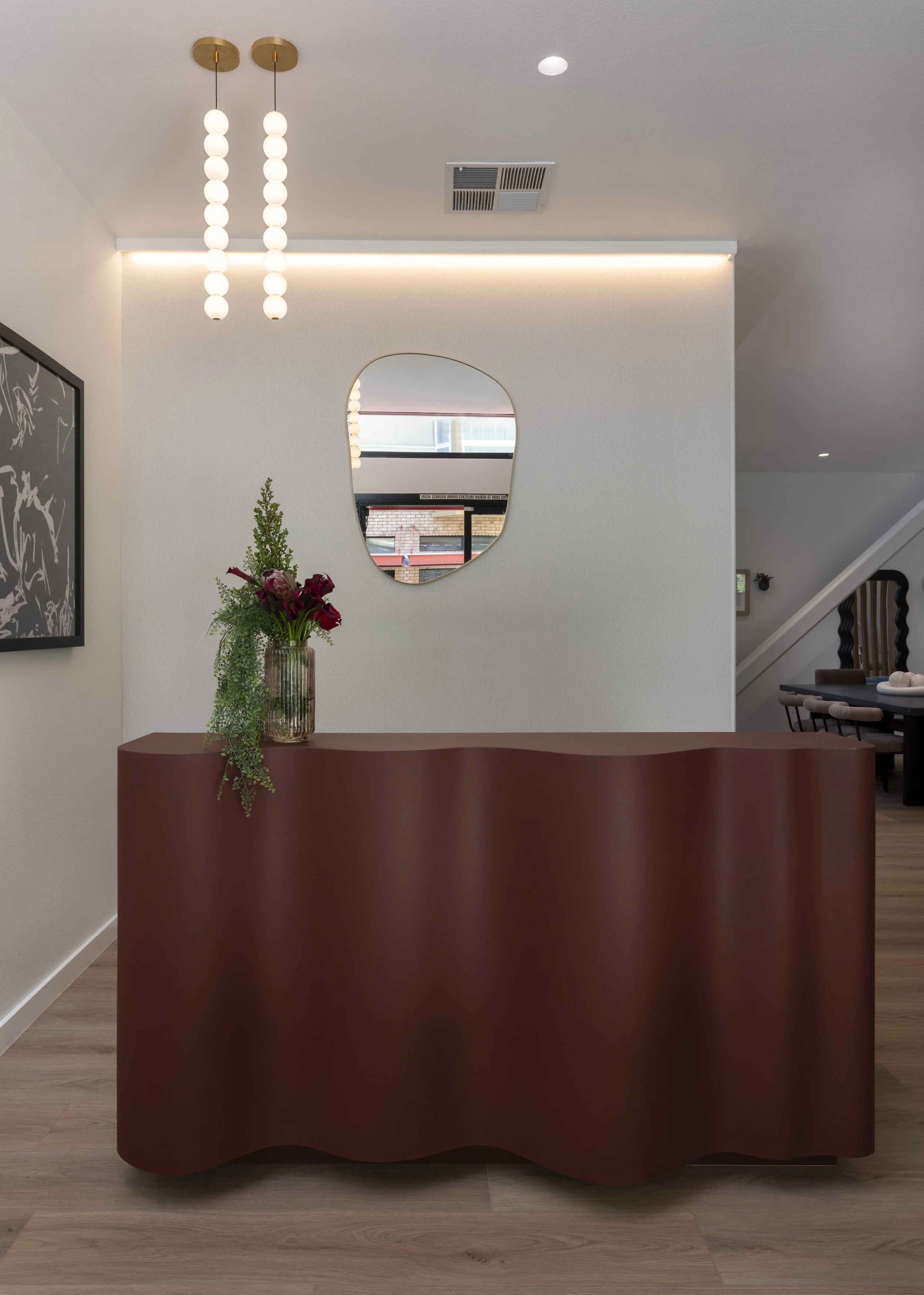 A modern interior with a red, wavy-front console table holding a floral arrangement with red and green foliage, a white wall with an abstract mirror, a black and white framed artwork, and contemporary lighting fixtures.