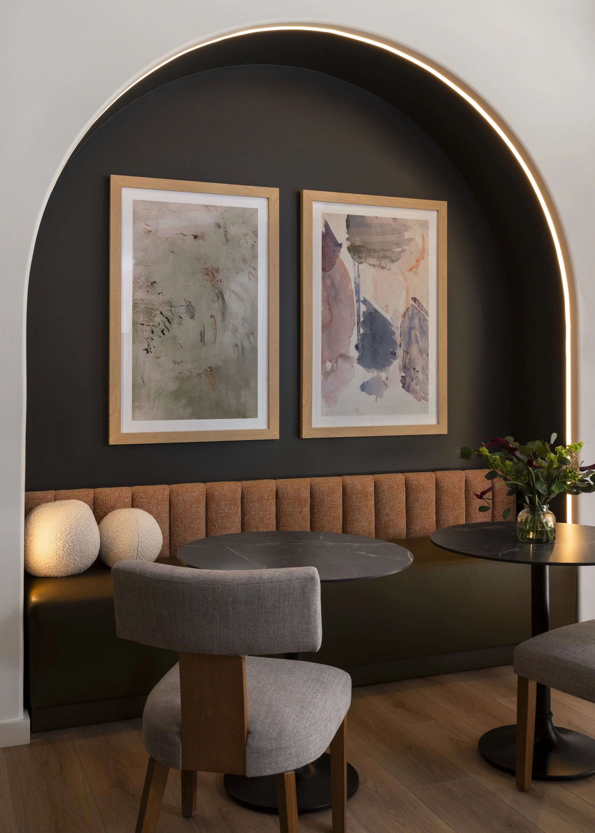 Interior of a modern dining area with two abstract watercolor paintings framed in wood on a dark wall, a curved upholstered banquette, a round black marble table, a gray upholstered chair, and a small round table with a glass vase of dark red and gre
