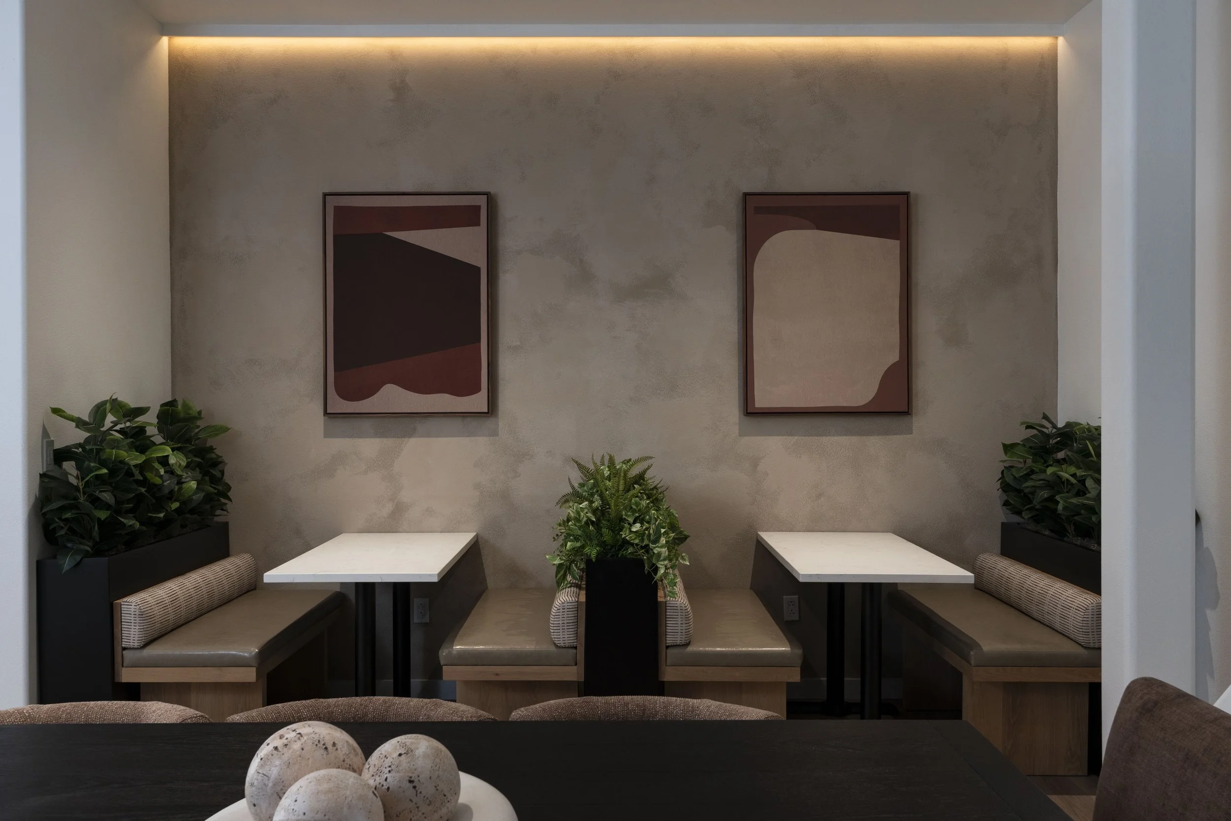 A cozy dining nook with beige upholstered bench seating, white tabletops, and green plants in black planters against a textured gray wall with framed abstract art, warm ambient lighting at the ceiling, and a dark wooden table with decorative vases in