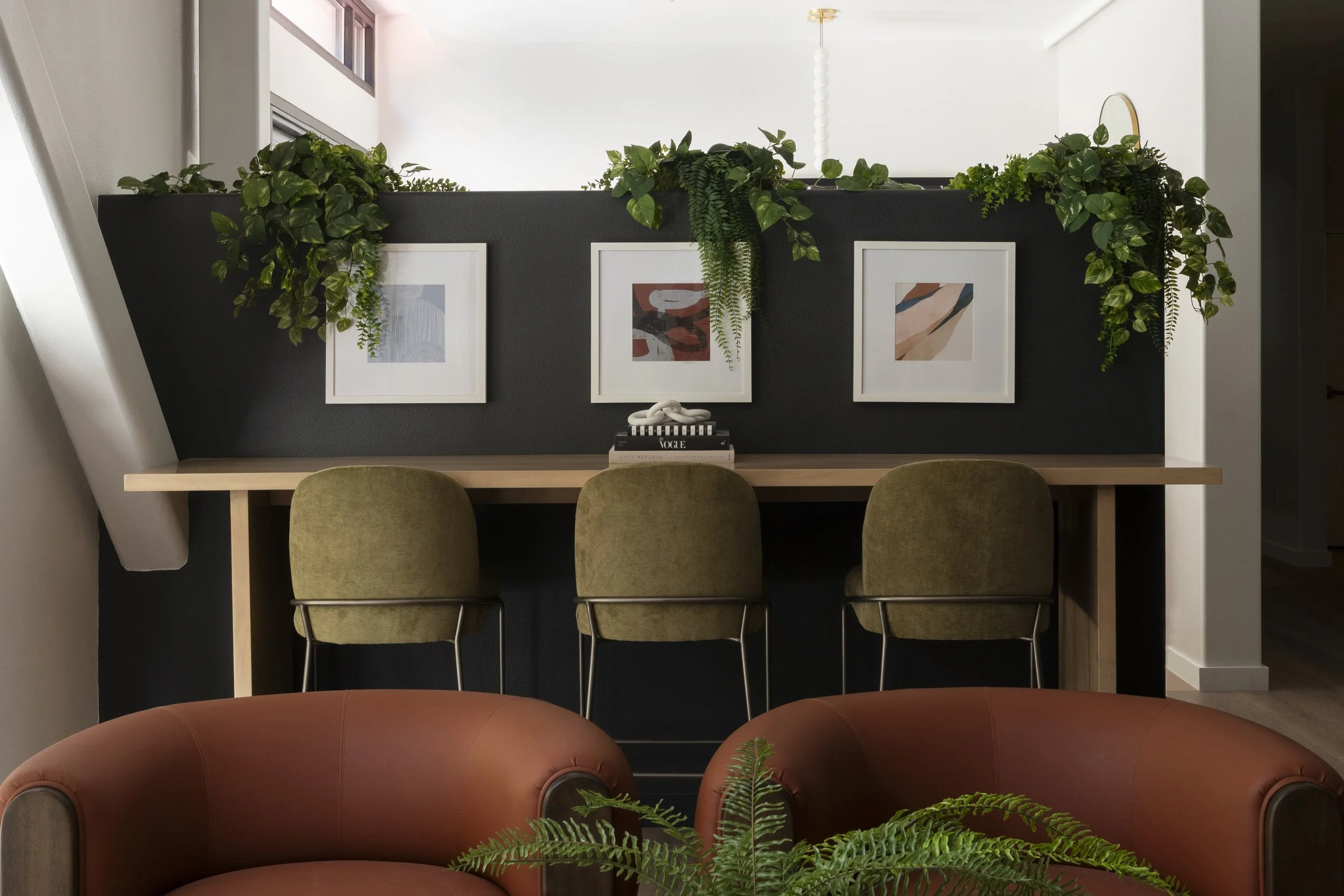A cozy living space with a black accent wall, three framed abstract art pieces, hanging plants, a wooden desk with three green upholstered chairs, and a reddish-brown sofa with a green fern in front.