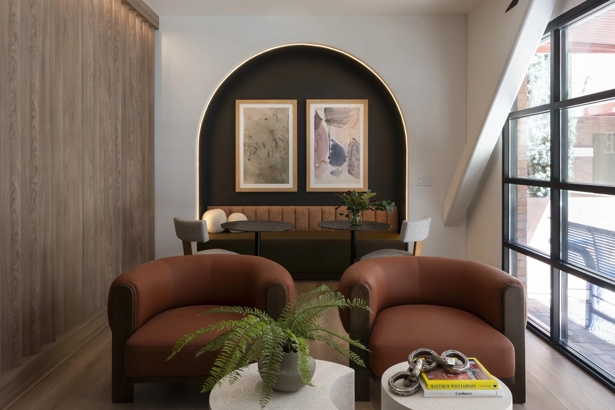 Modern living room with two brown leather armchairs, a white coffee table with a plant and a stack of books, and a built-in window seat with a small table and two chairs. Wall art hangs on a black accent wall with an illuminated arch.