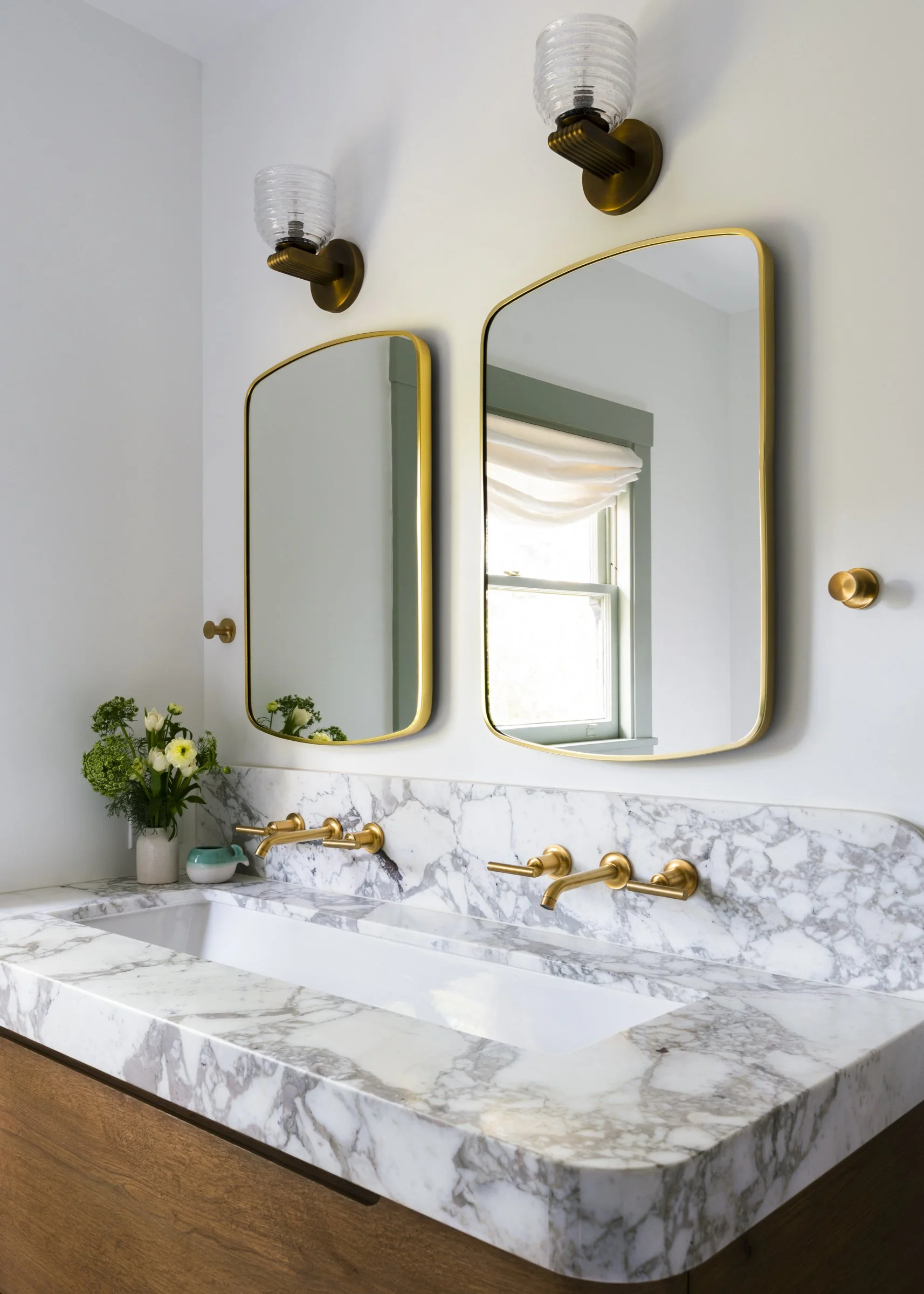 A modern bathroom vanity with a white marble countertop, two gold faucets, two mirrors with gold frames, wall-mounted light fixtures, a small vase with flowers, and a window with a white shade.