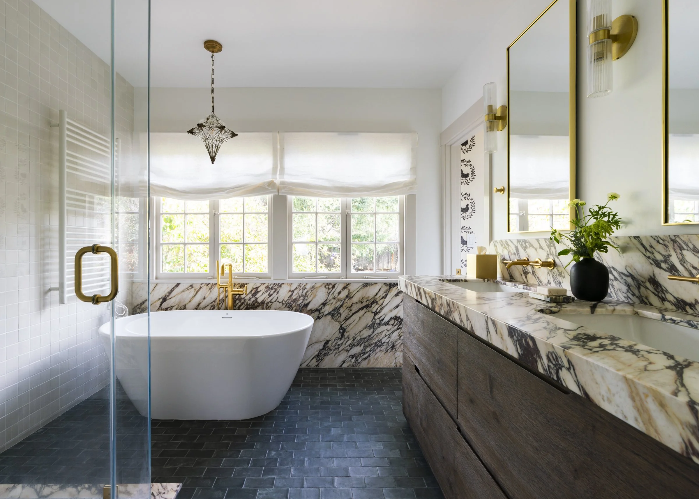 Modern bathroom with a white bathtub, a wooden vanity with a marble countertop, and gold fixtures. Large window with blinds, black floor tiles, and a glass shower door. Decor includes a potted plant and mirrors with gold frames.
