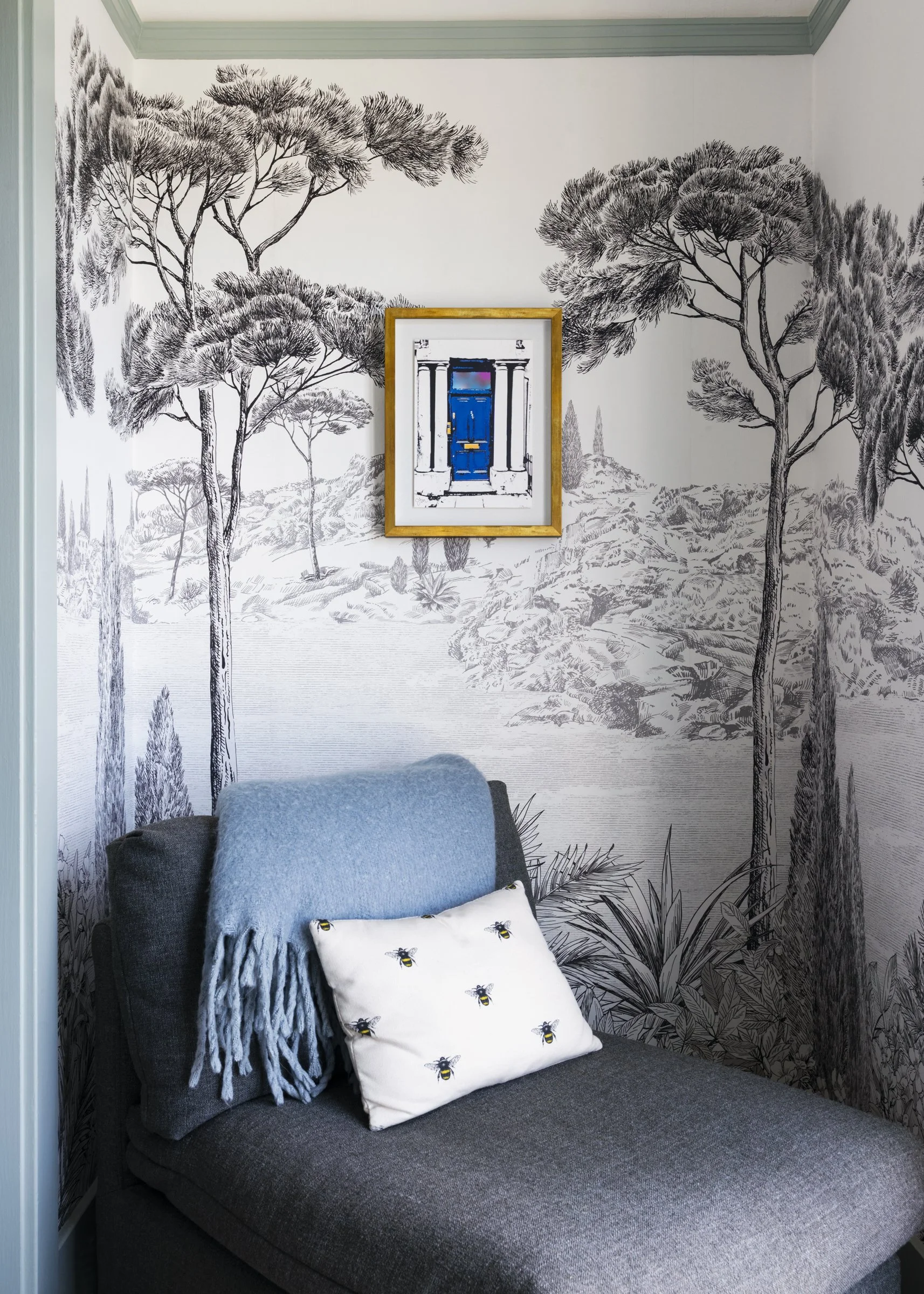 A corner of a room with a wall mural of black and white trees and landscape, a framed picture of a blue door, a gray armchair with a blue blanket, and a decorative pillow with bee patterns.