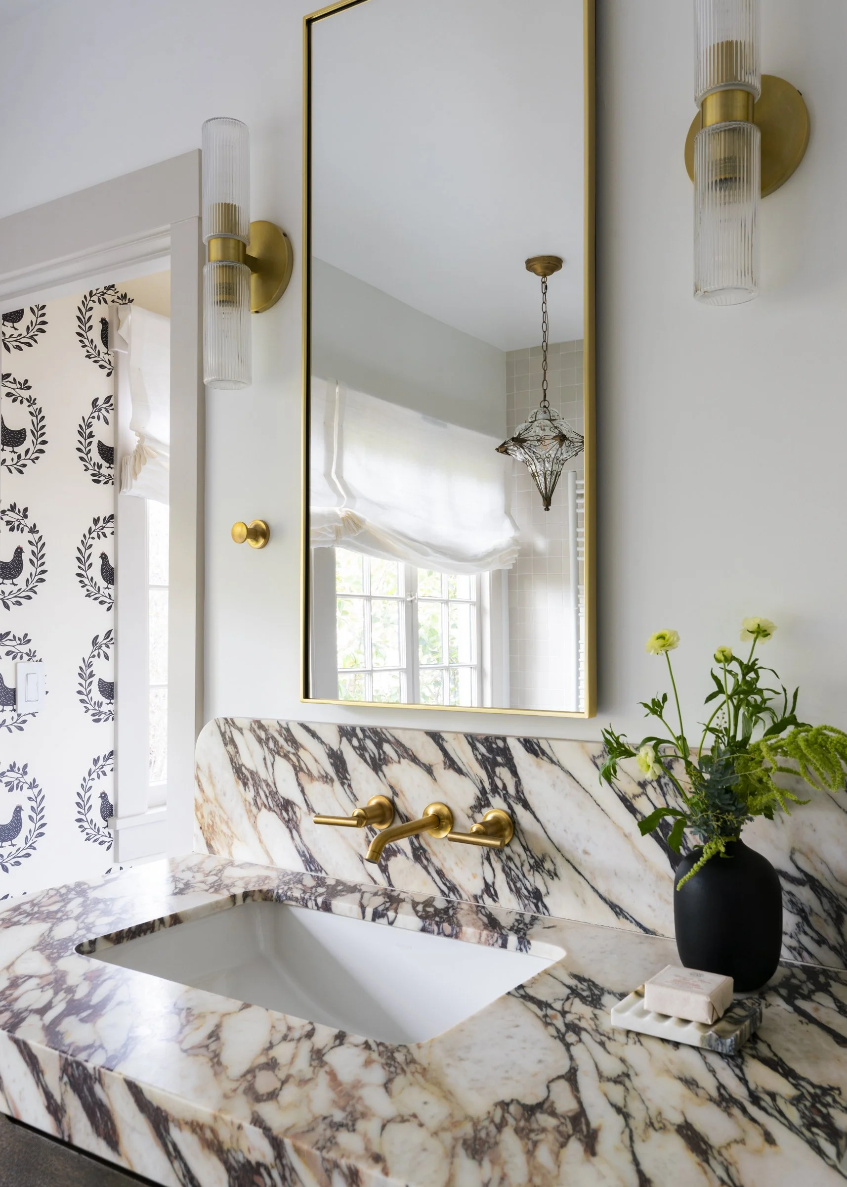 A bathroom vanity with a marble countertop and backsplash, gold fixtures, a black vase with green and yellow flowers, a mirror, and modern wall sconces, with a window and light-colored walls.