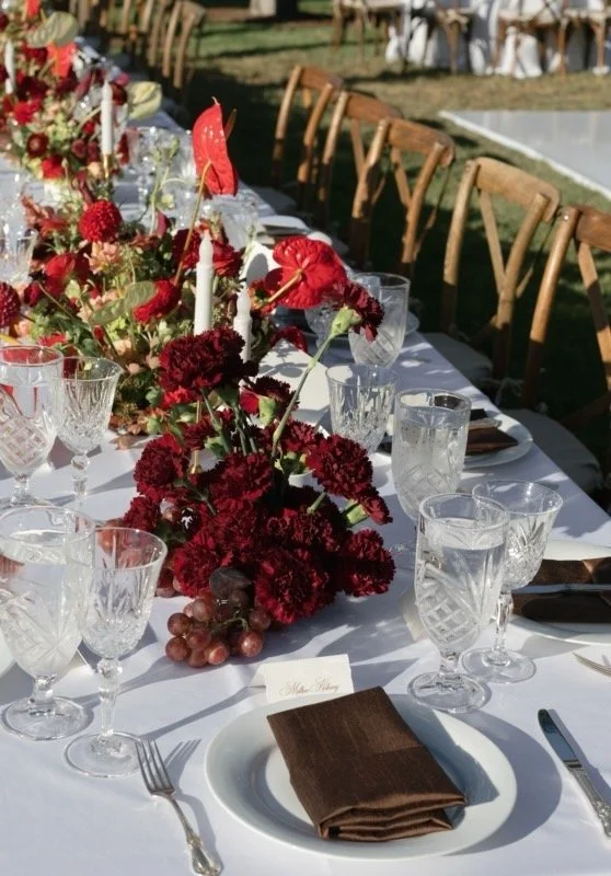 A long dining table set outdoors with white tablecloths, floral centerpieces in red and burgundy, glassware, plates, and folded brown napkins, surrounded by wooden chairs.