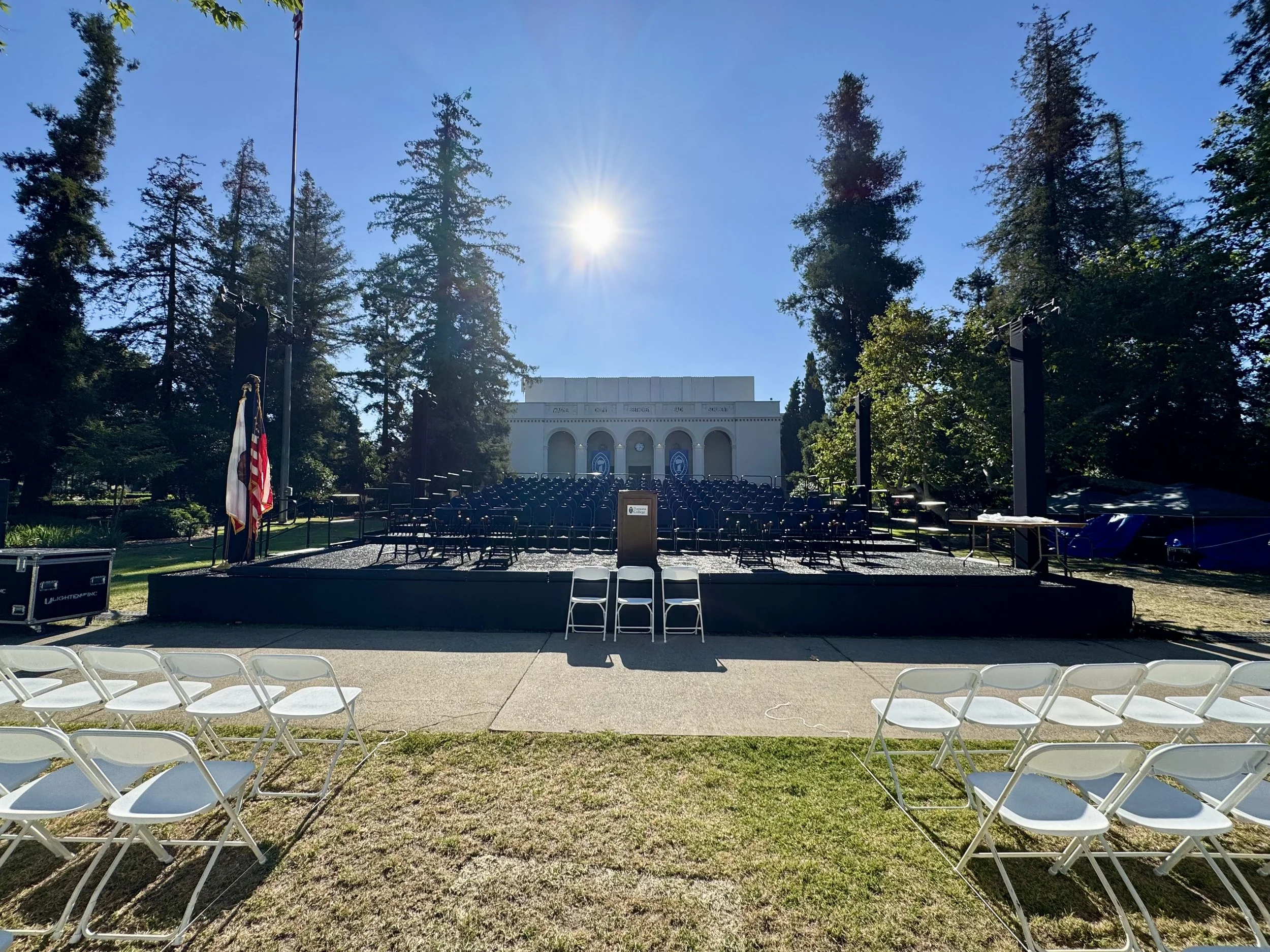 Outdoor stage setup for a ceremony with chairs arranged facing the stage, American flag, surrounded by tall trees, and a historic building in the background on a sunny day.