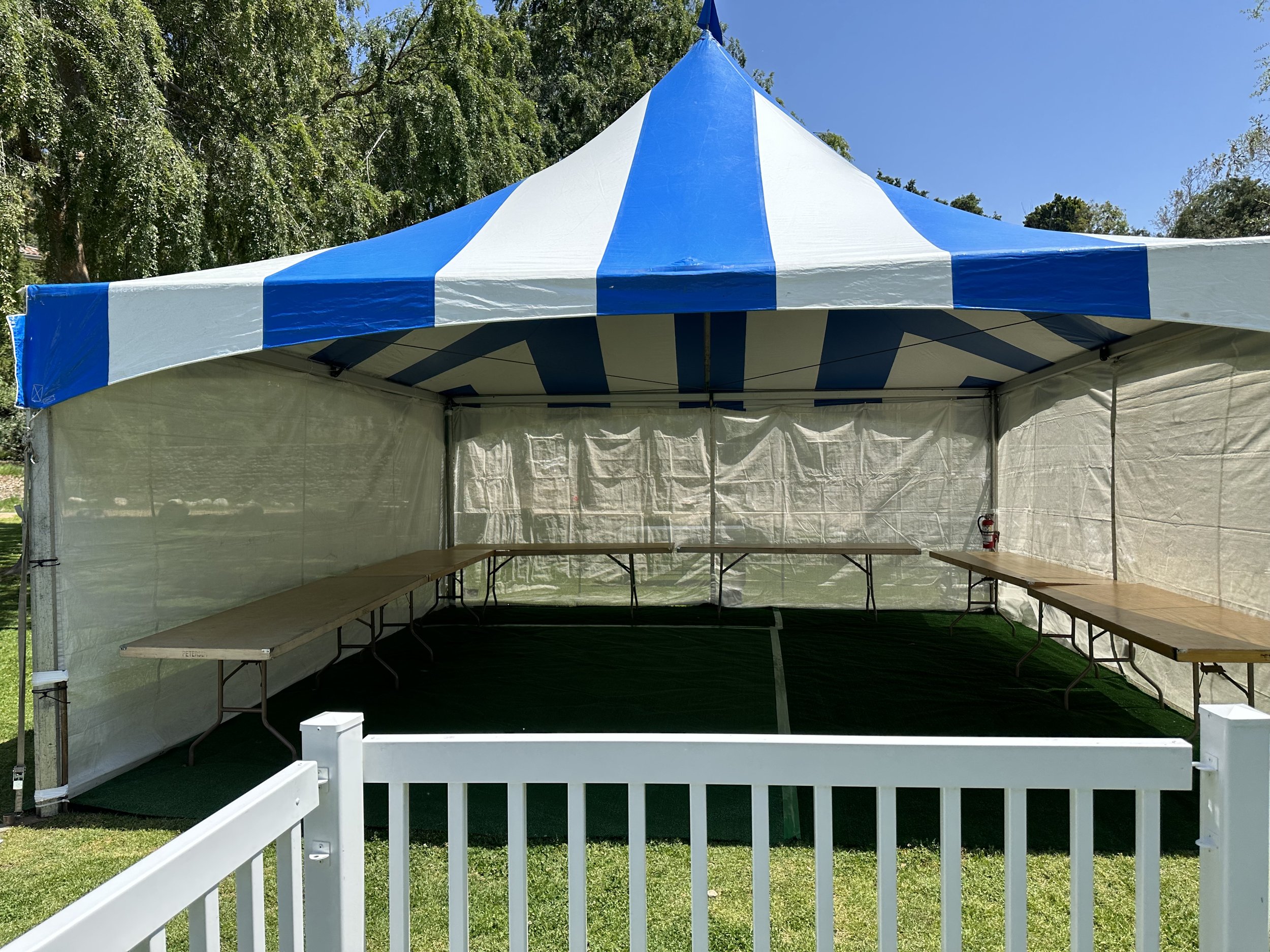 Empty blue and white striped tent with tables, outdoors on grass, enclosed by a white fence, with trees in the background.