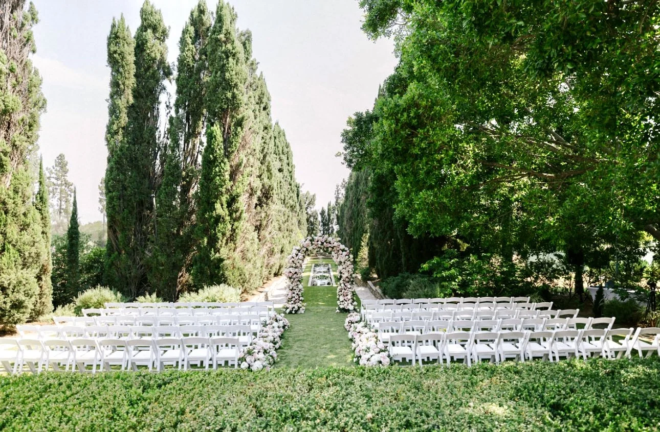 Outdoor wedding ceremony setup with rows of white chairs, a floral arch, and lush green trees and bushes.