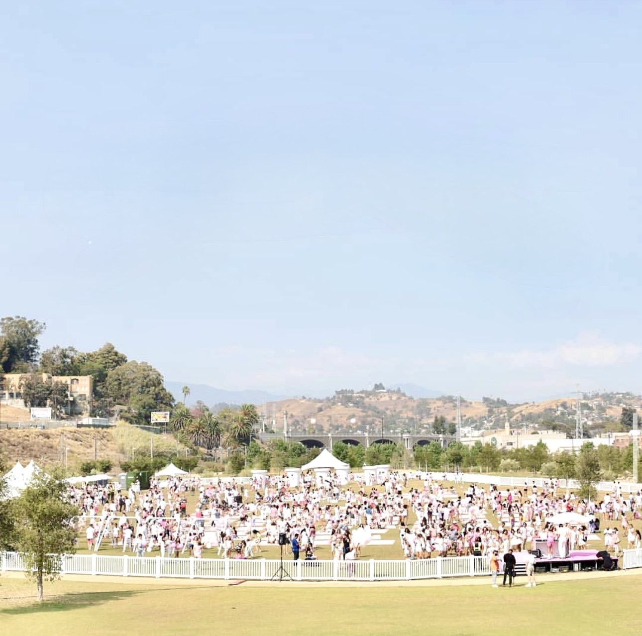 Crowd of people dressed in pink gathering in an open field during daytime, with tents and a fence in the foreground, and hills and a bridge in the background.
