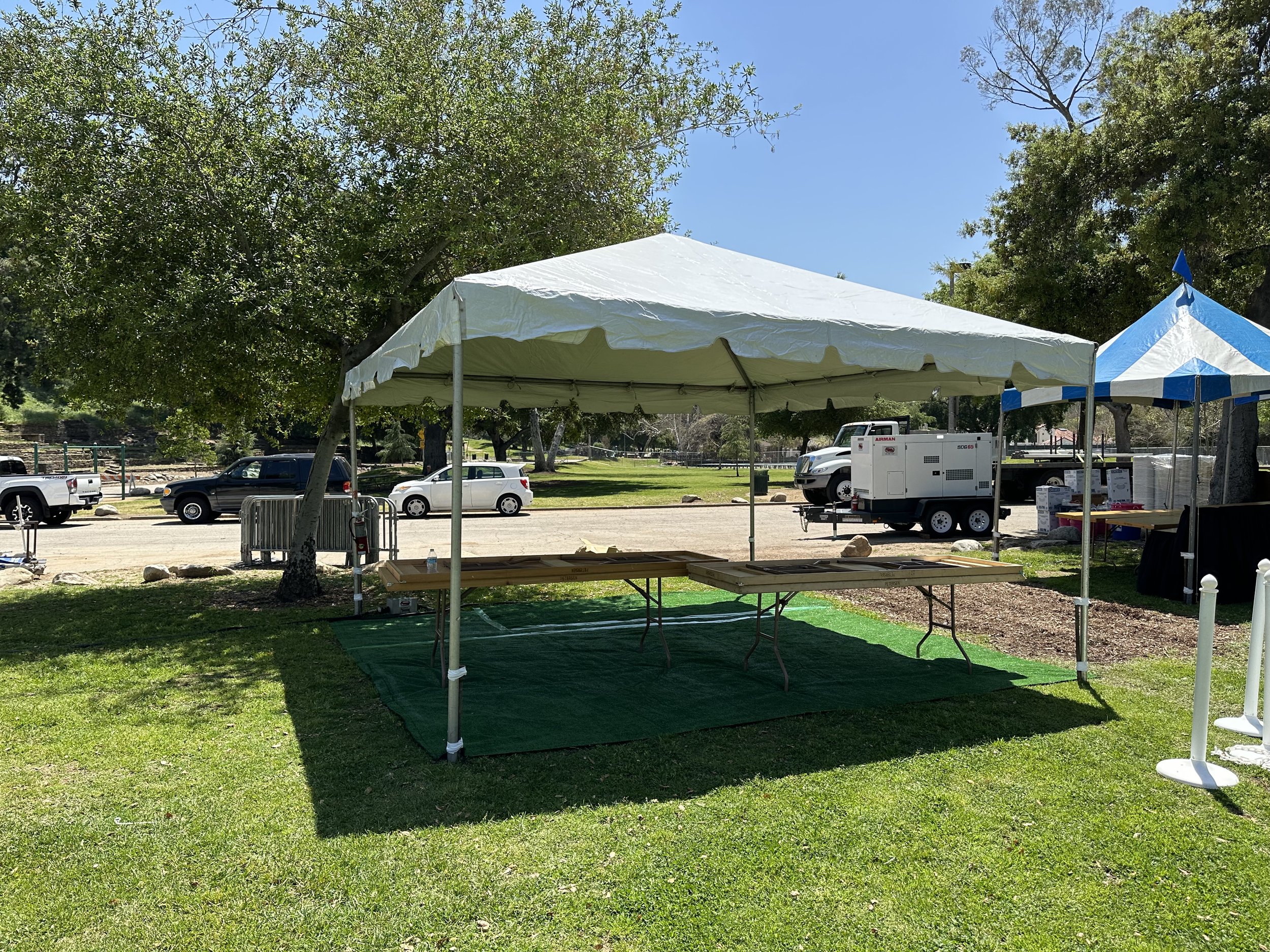 Outdoor event setup with a white canopy tent over tables, a nearby generator, vehicles, and grassy surroundings.