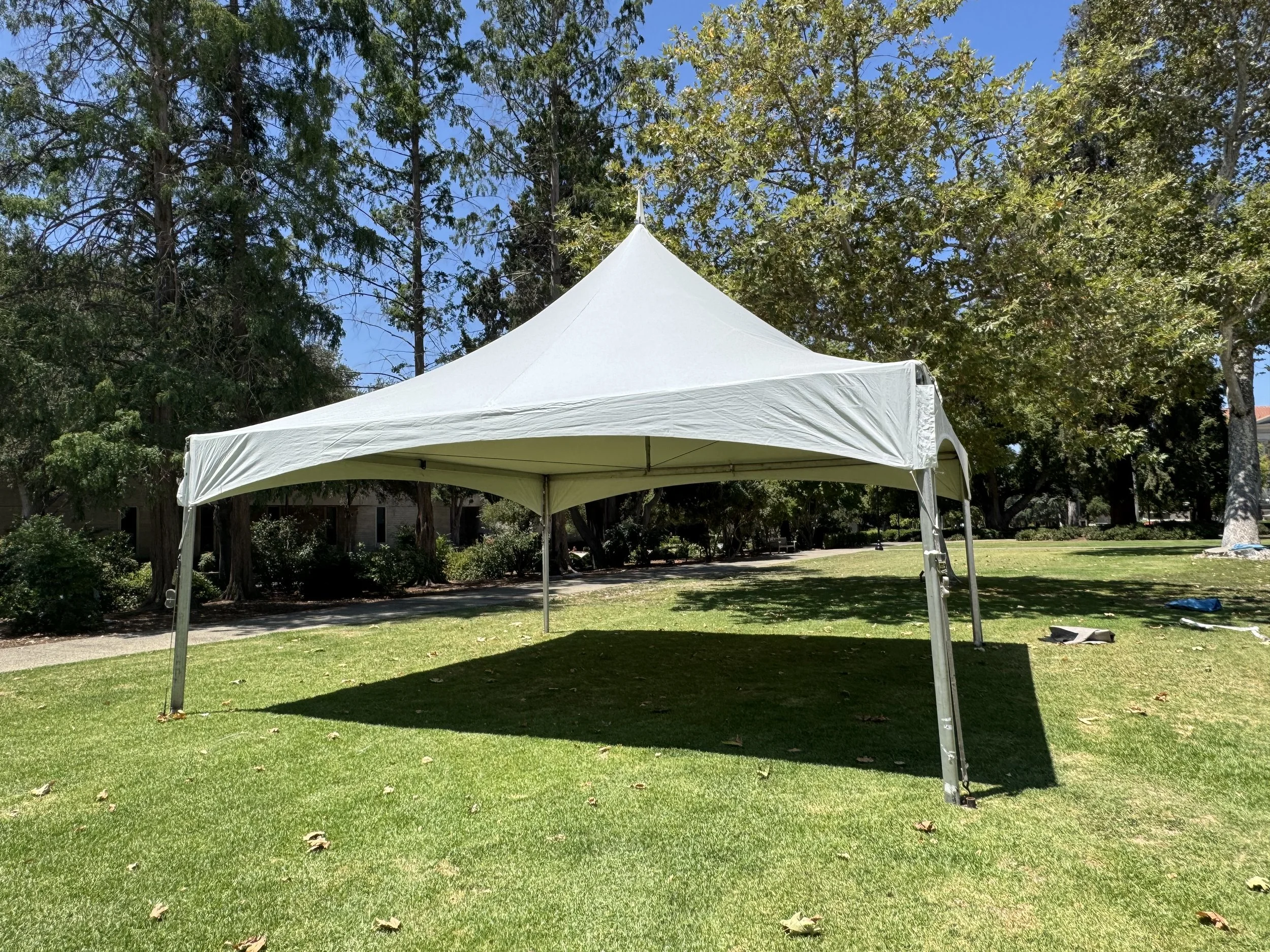 A white outdoor canopy tent set up on a grassy lawn with trees and a walking path in the background on a sunny day.