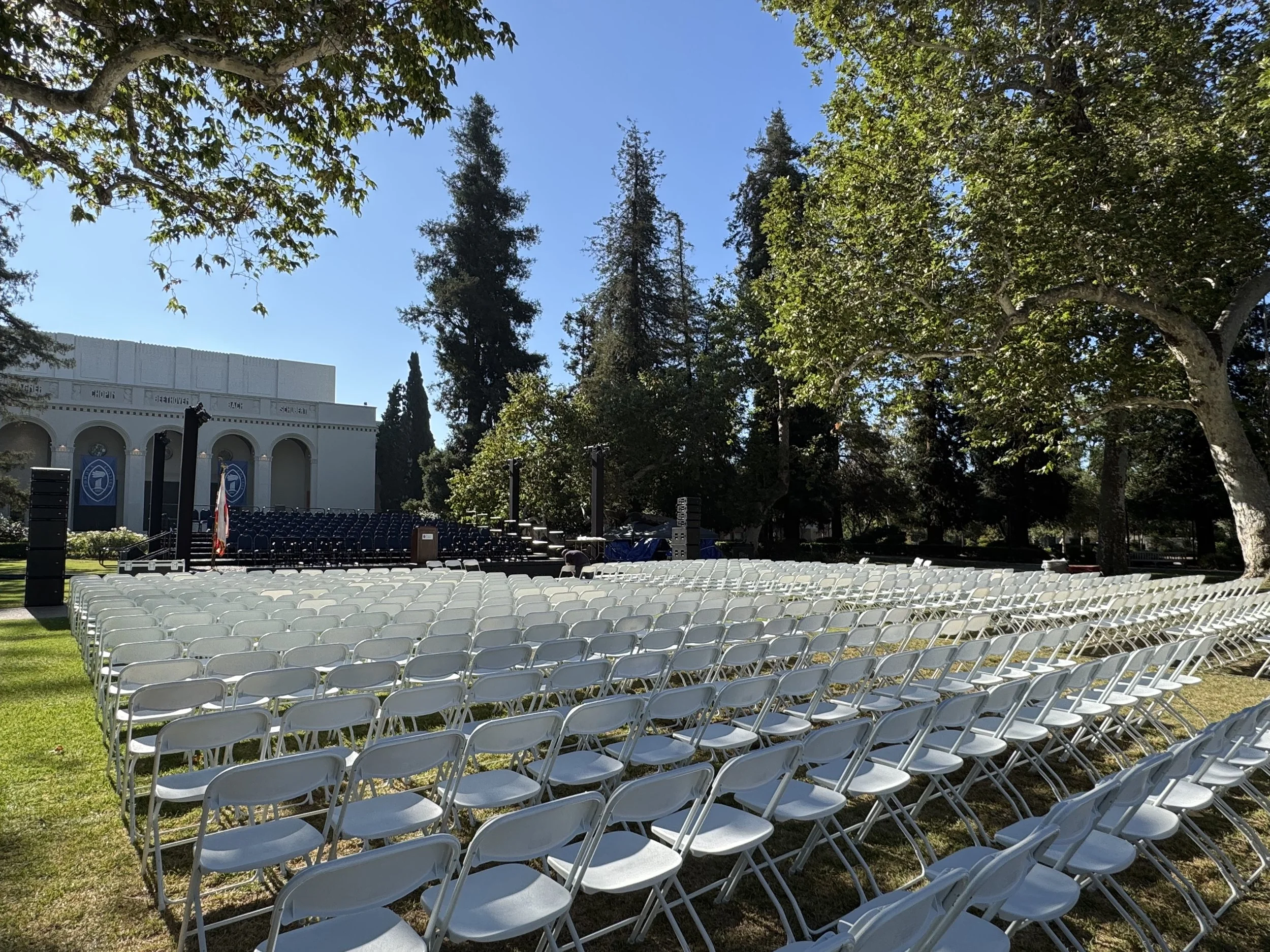 An outdoor stage setup with rows of white folding chairs on a grassy lawn, surrounded by trees and a large building in the background, under a clear blue sky.