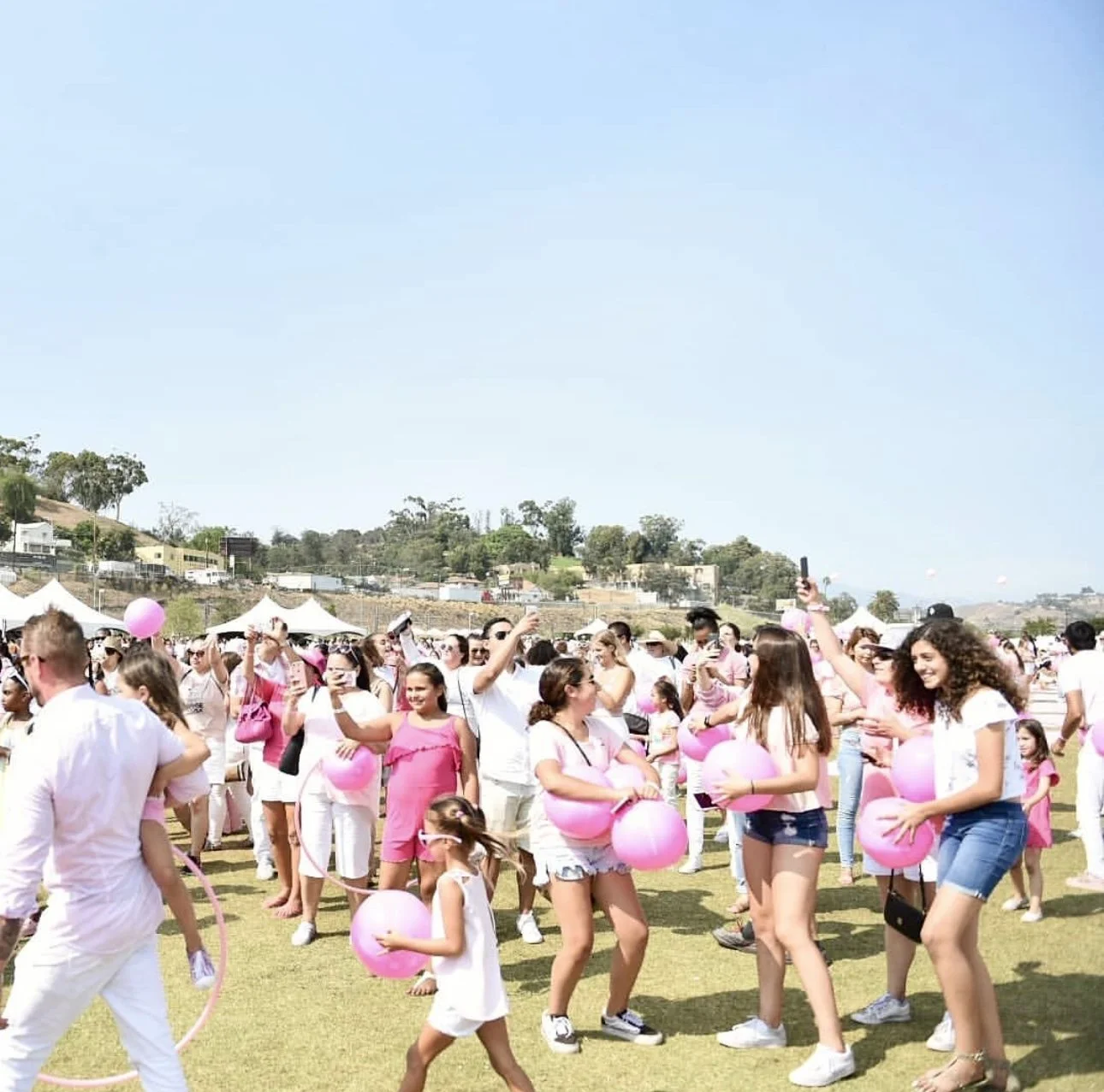 People gathered outdoors on a grassy field participating in a color run event, holding pink balloons, with some dancing and others taking photos under a clear blue sky.