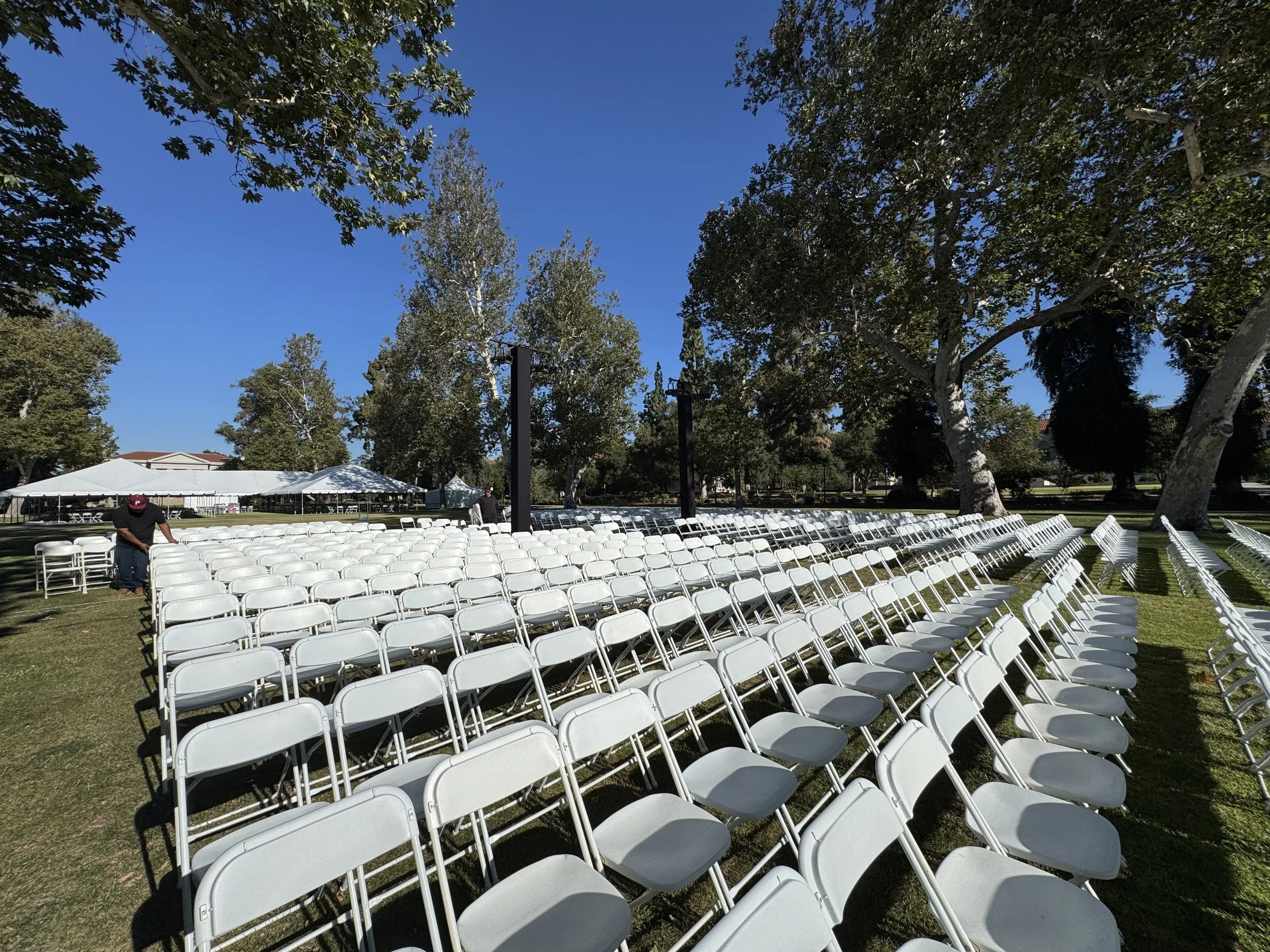 Rows of white folding chairs set up outdoors under a clear blue sky with trees and tents in the background, likely for an outdoor event or ceremony.