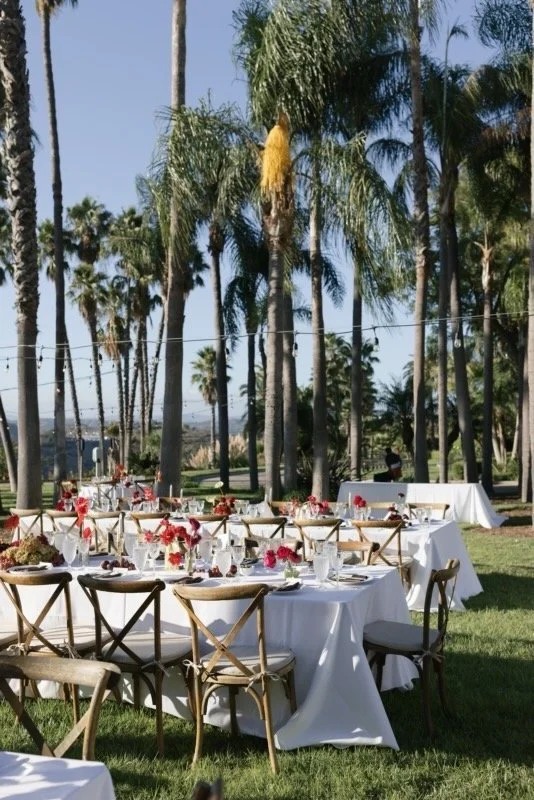 Outdoor event setup with round tables covered in white tablecloths, decorated with pink and red floral arrangements, set with glassware and silverware, surrounded by wooden chairs, under a grove of tall palm trees on a sunny day.