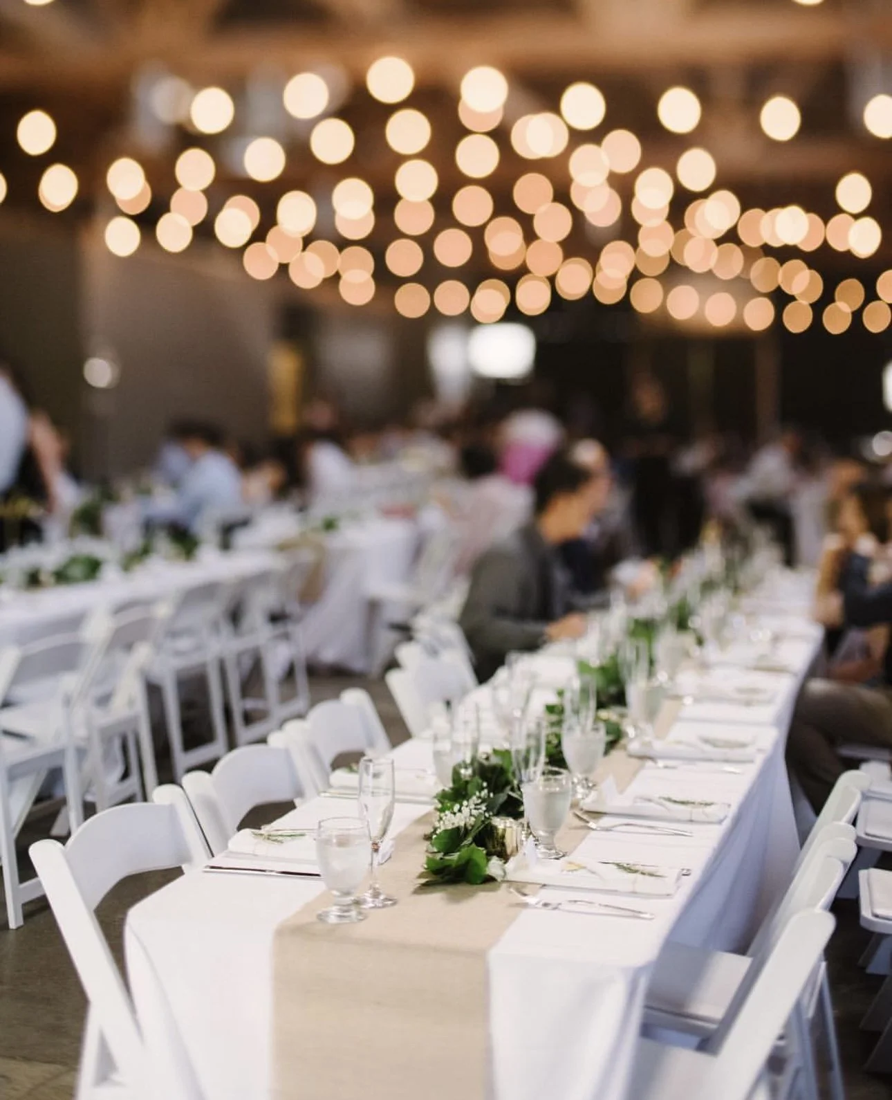 A long banquet table decorated for a formal event with white chairs, greenery, and water glasses, under string lights with blurred guests in the background.