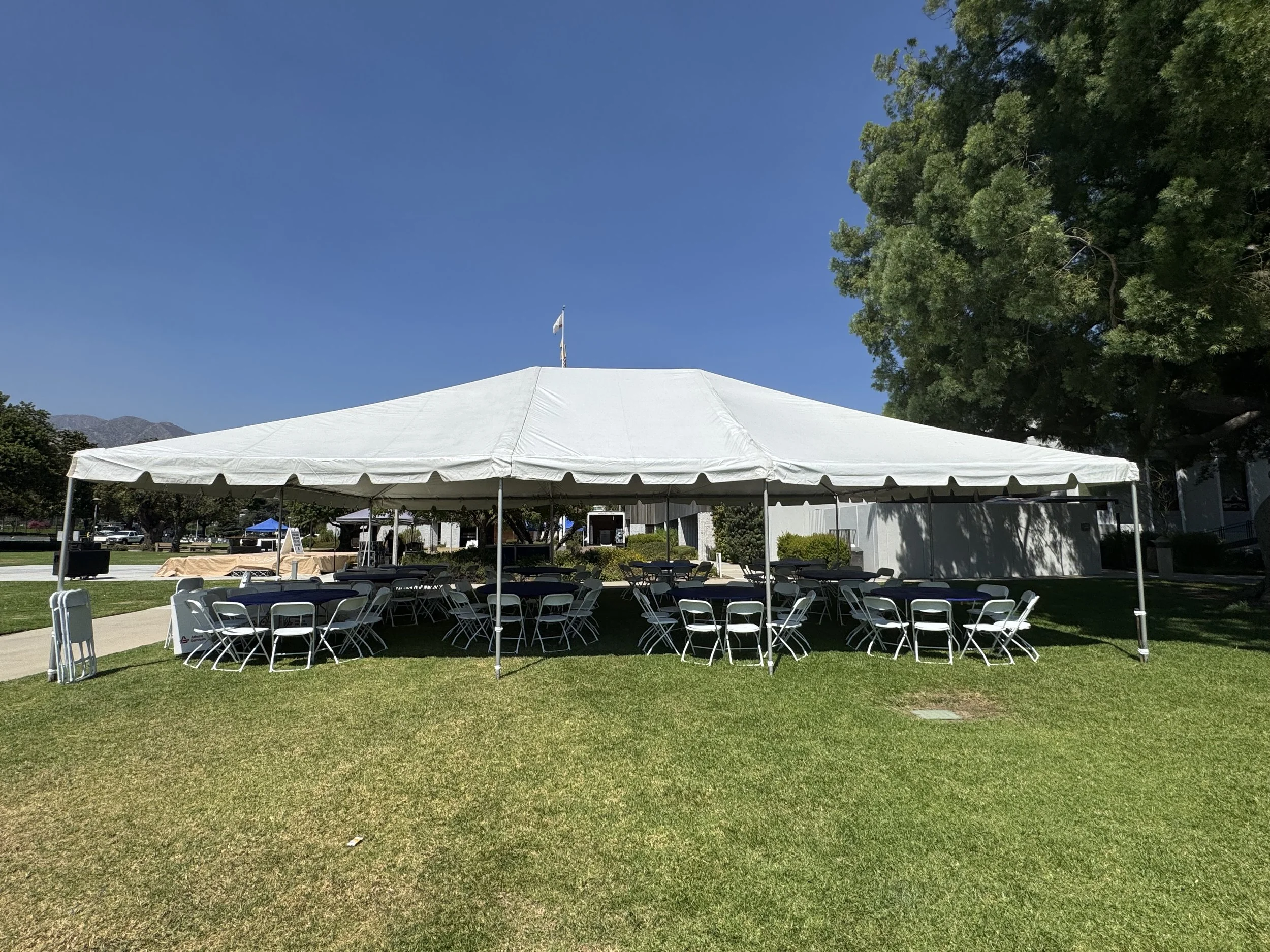 Large white event tent set up on grassy area with tables and chairs underneath, clear blue sky, trees in background.